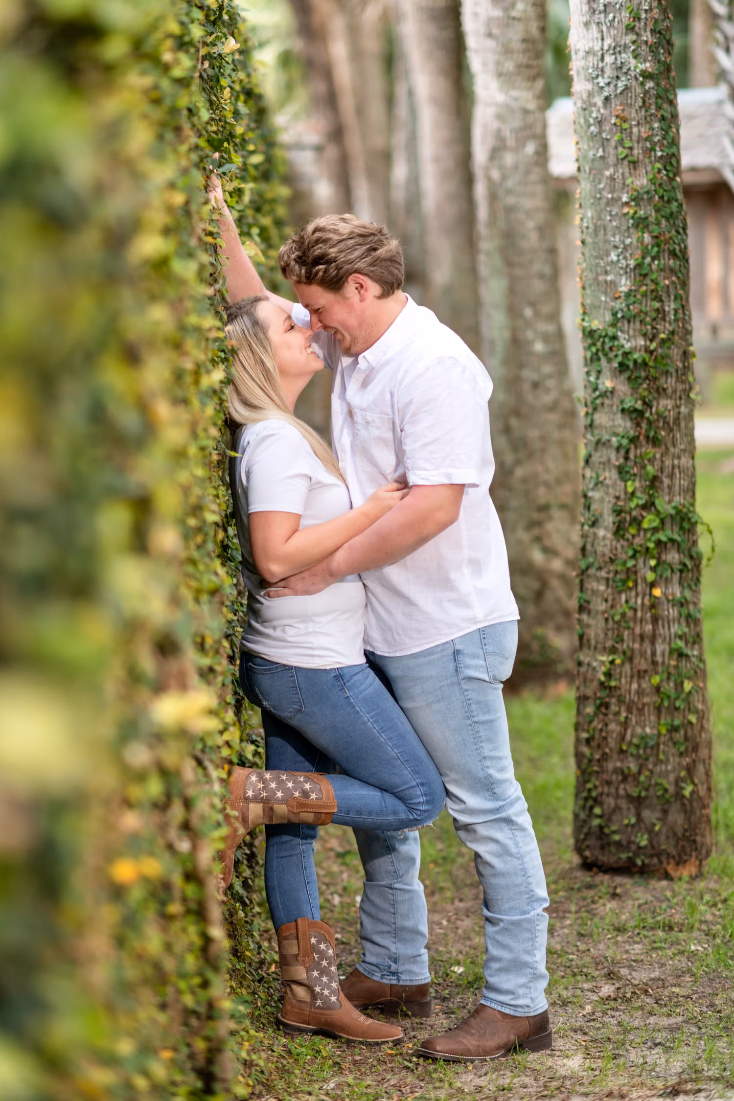Woman leaning against an ivy-covered wall as her fiancé leans in for a kiss at Huntington Beach State Park