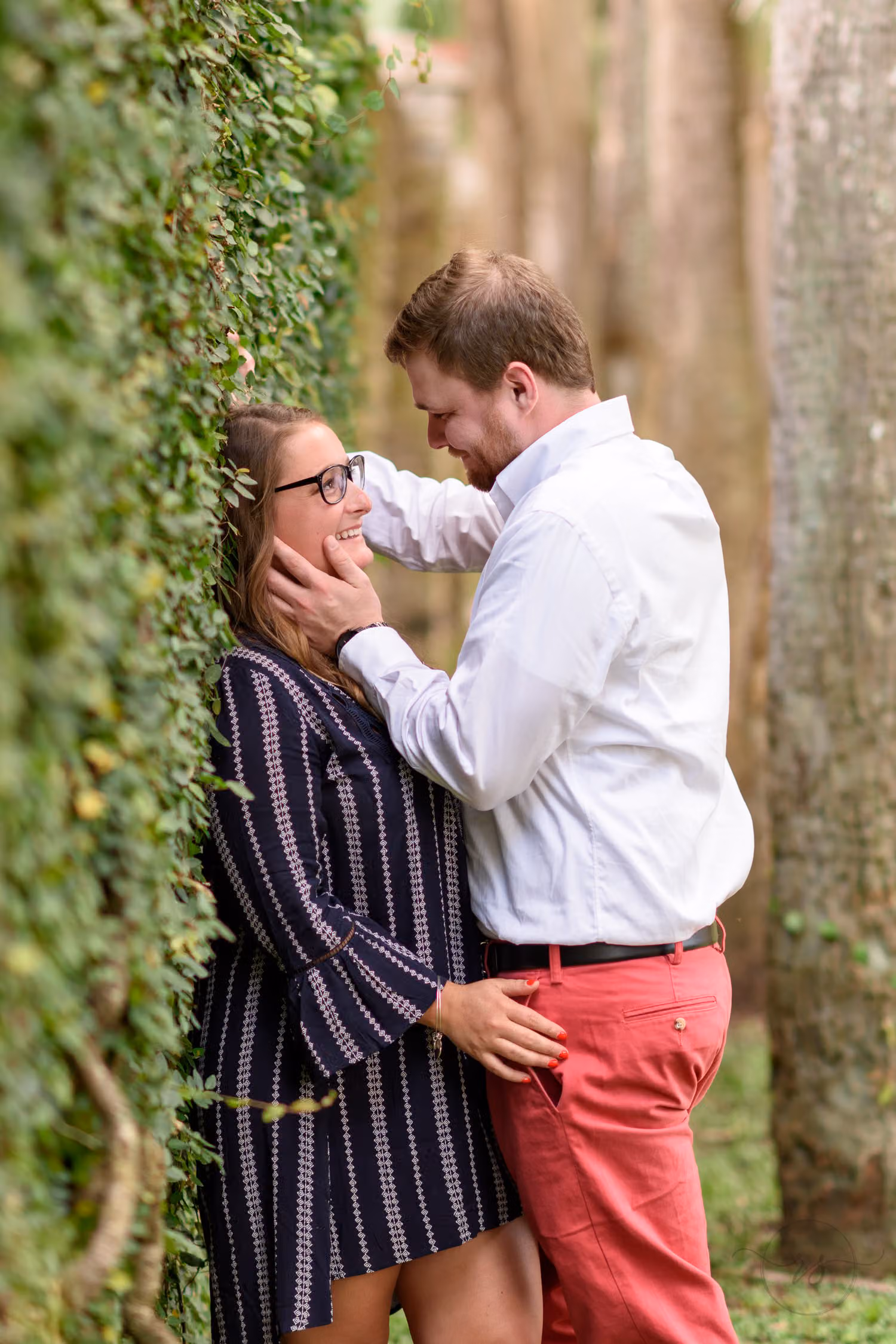 Man touching his fiancée's face as she leans back against an ivy-covered wall beside tall trees