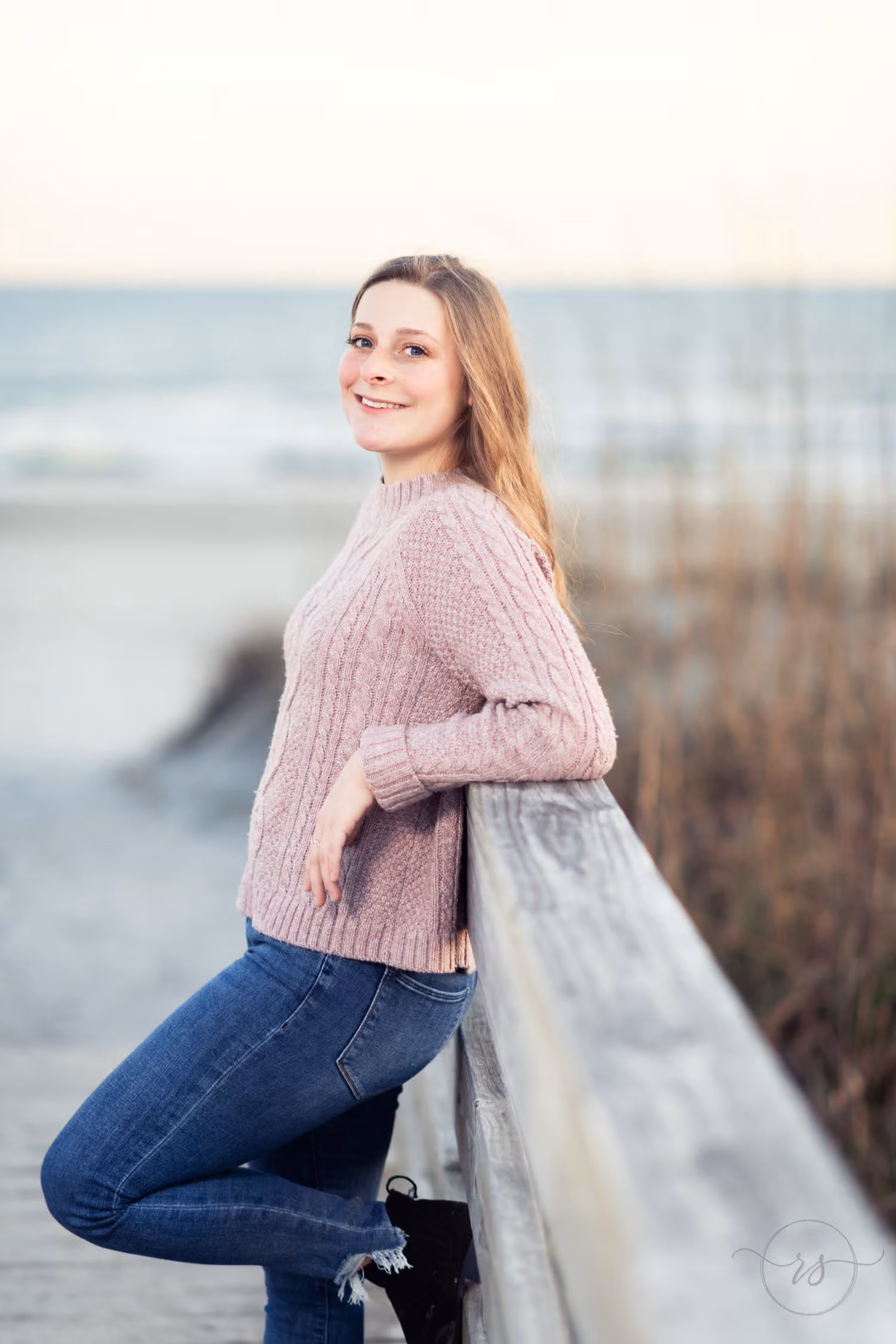 Senior in a pink sweater leaning on the boardwalk railing with the beach behind her at Huntington Beach State Park