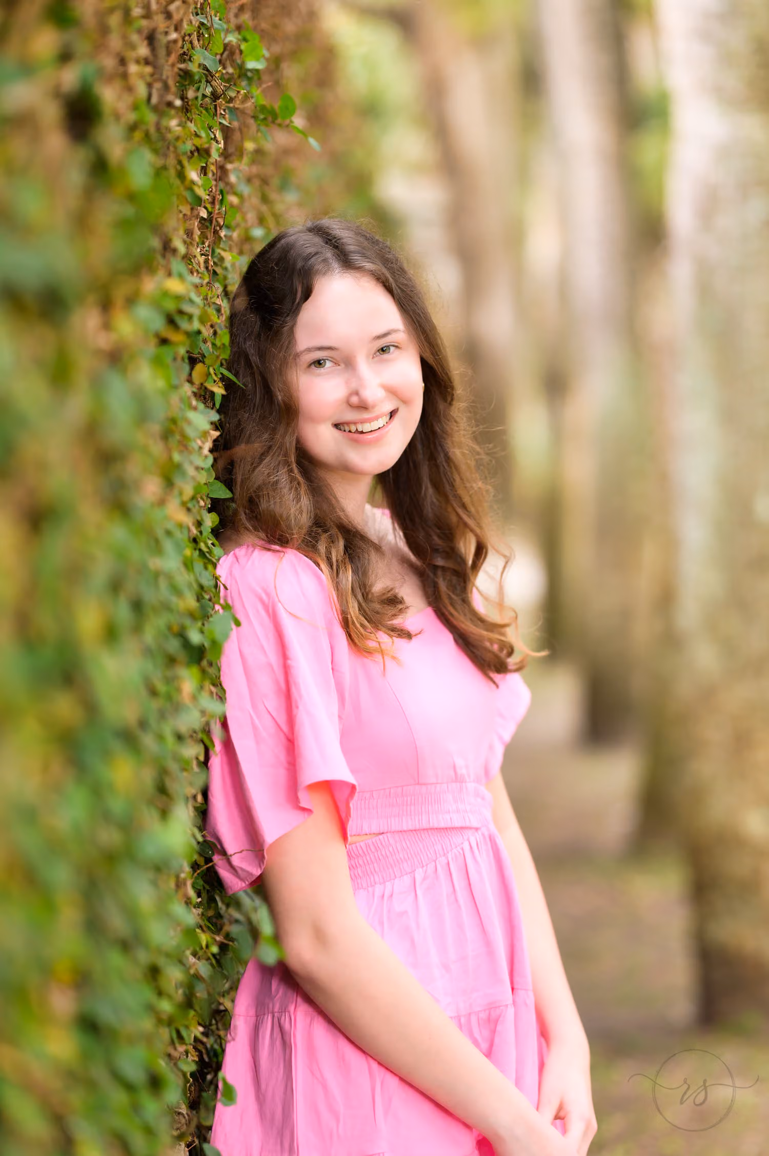 Senior in a pink dress leaning against an ivy-covered wall lined with palm trunks at Huntington Beach State Park