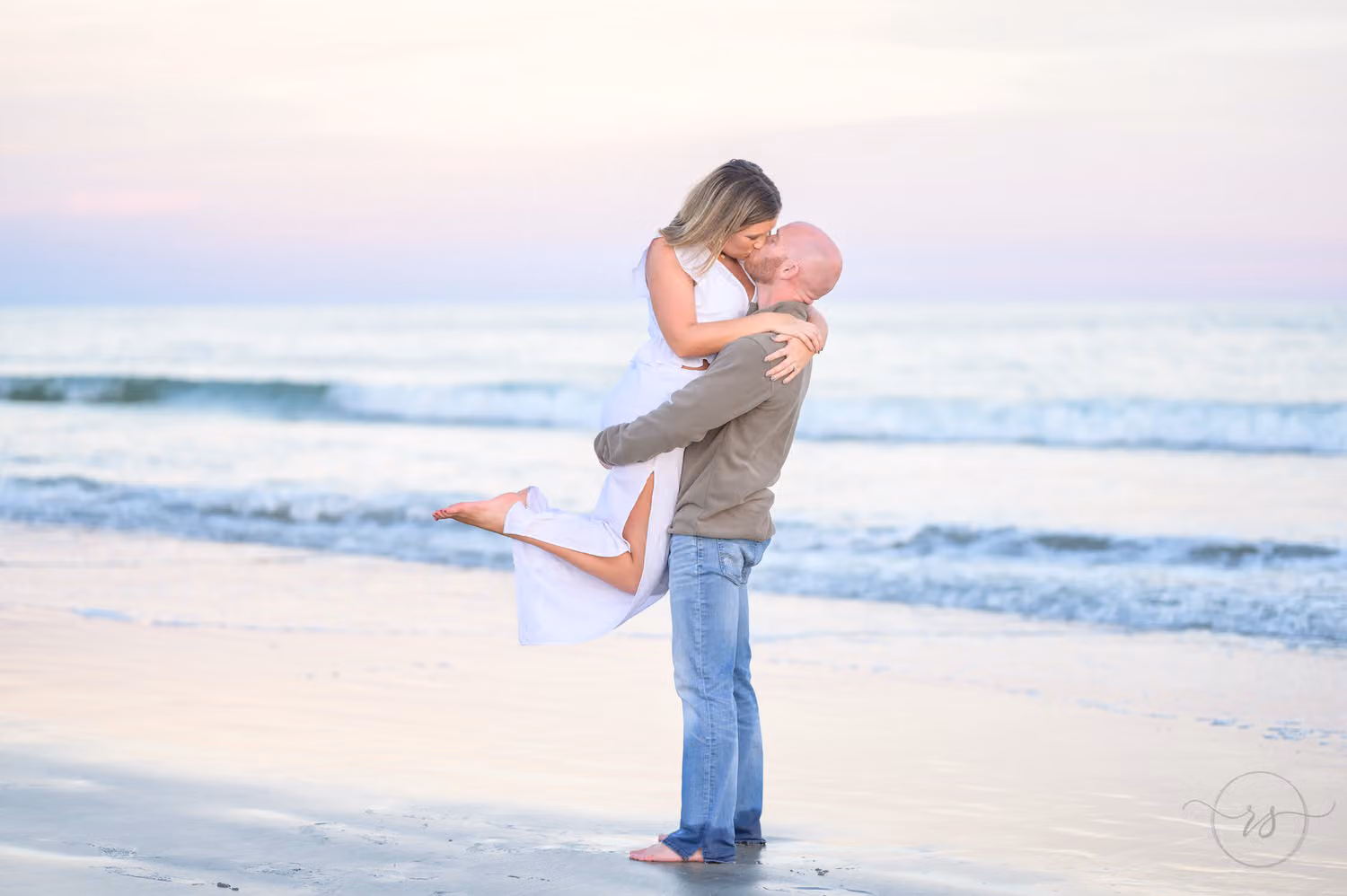 Man lifting his fiancée into the air for a kiss as she kicks her feet up by the ocean at Huntington Beach State Park
