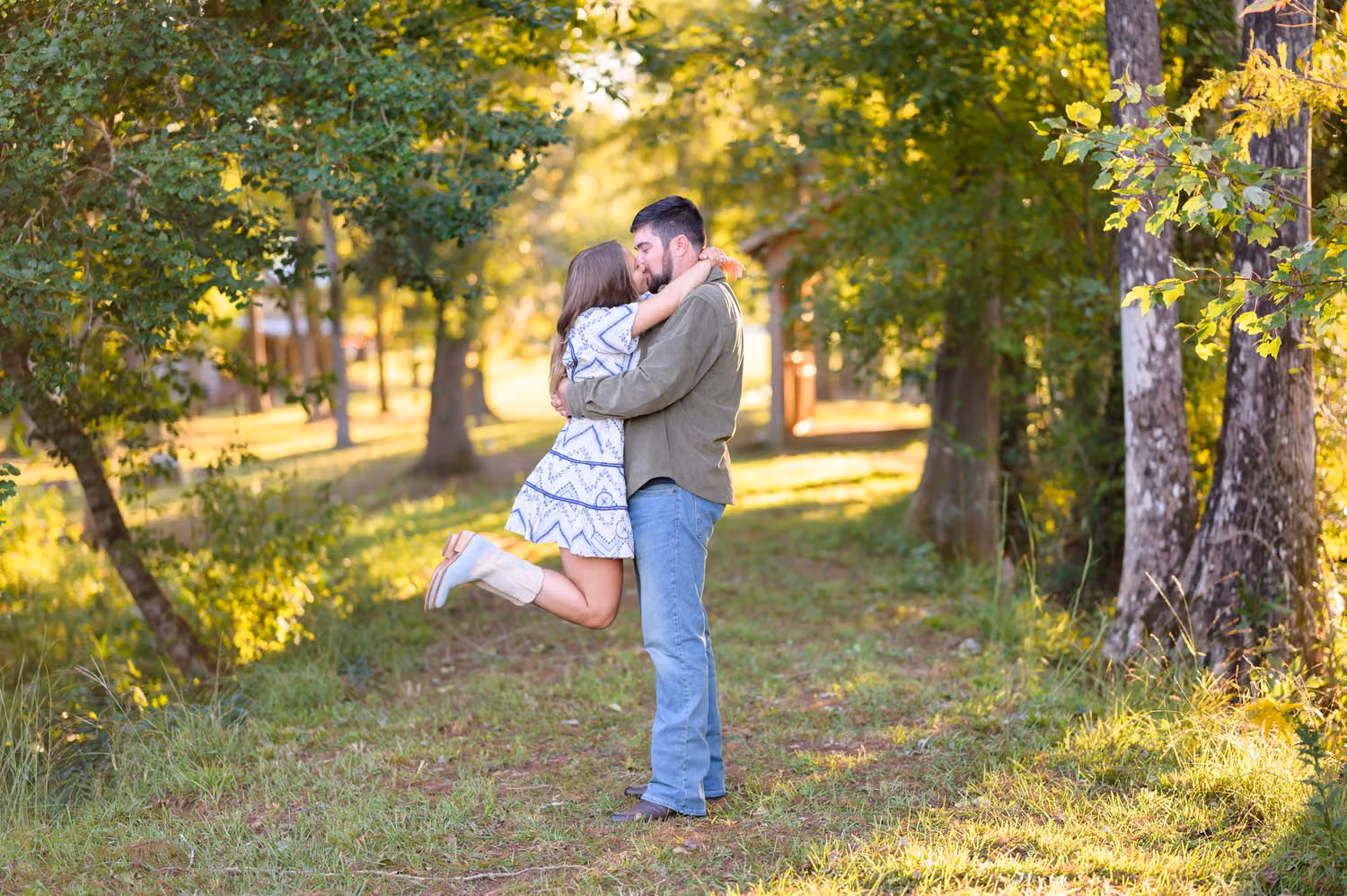 Woman in cowboy boots kicking up a heel as her fiancé lifts her for a kiss in the sunny woods at Pine Log Plantation