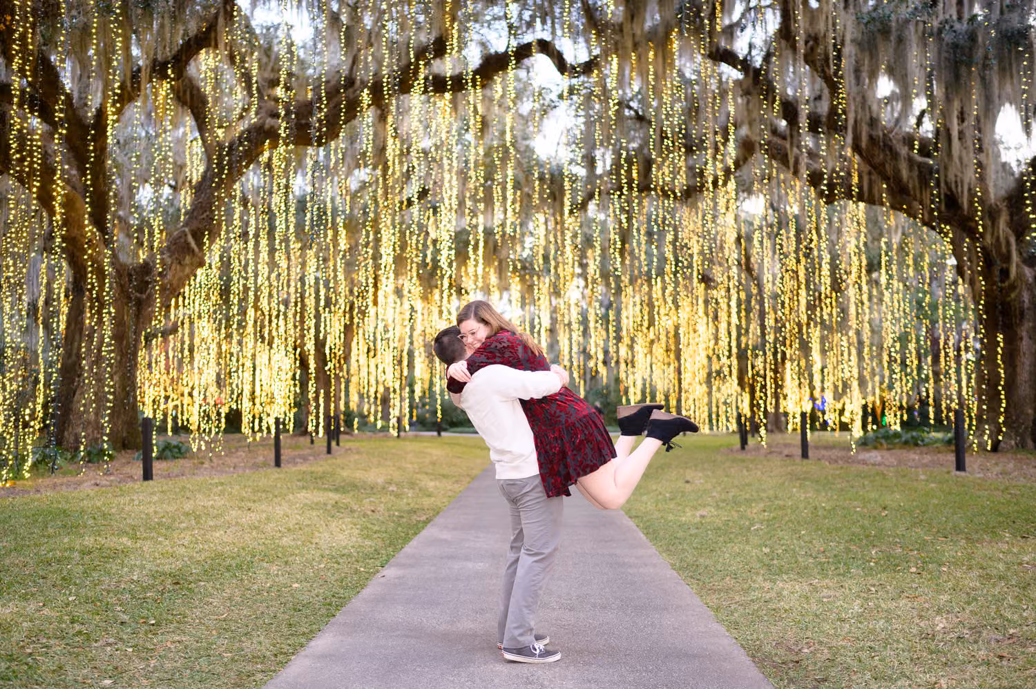 Man lifting his fiancée on a path under live oaks hung with strands of glowing lights at Brookgreen Gardens