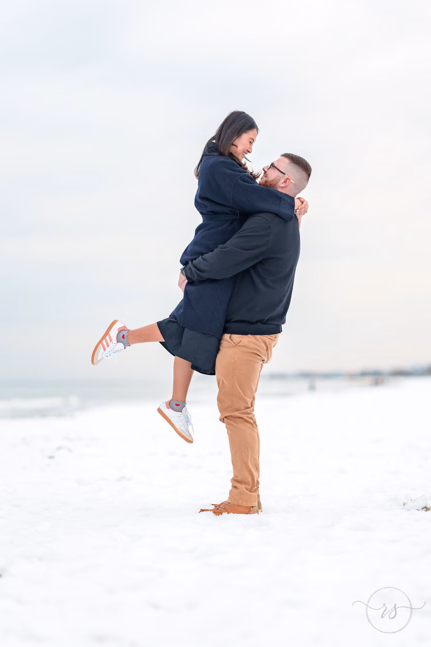 Woman in a long coat wrapping her arms around her fiancé as he lifts her on the snowy sand on Garden City Beach