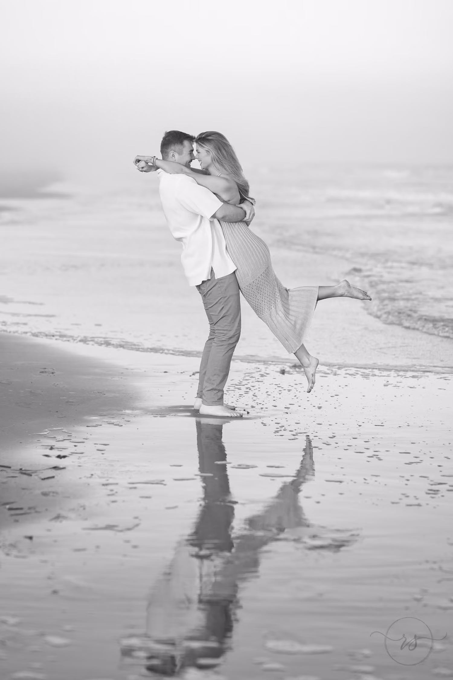 Black and white of a man lifting his fiancée on the wet sand with their reflection below at Huntington Beach State Park