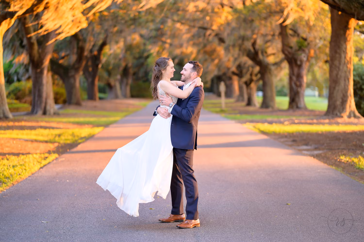 Groom lifting the bride into the air on a road lined with moss-covered oaks in golden light at Caledonia Golf & Fish Club