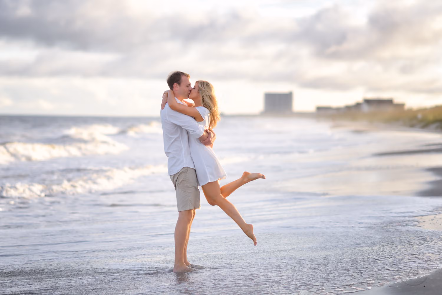Man lifting his fiancée off the sand for a kiss as she kicks up her foot by the surf at Myrtle Beach State Park