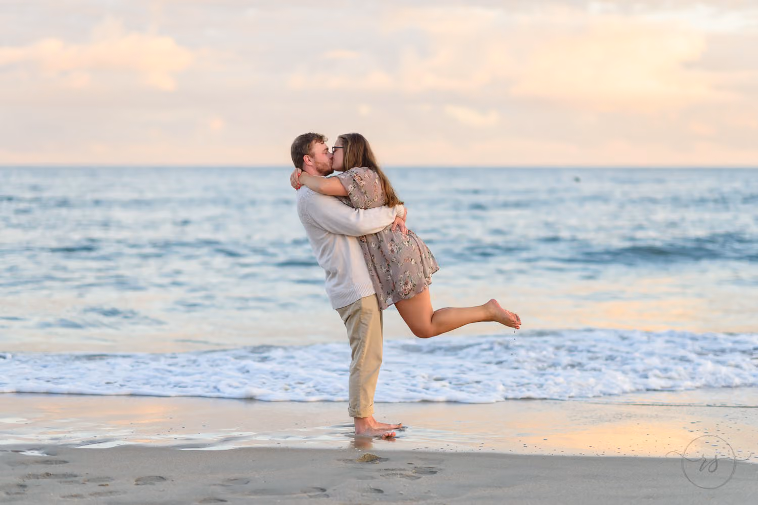 Man lifting his fiancée off her feet as she kicks up a leg at the edge of the ocean under a soft winter sky