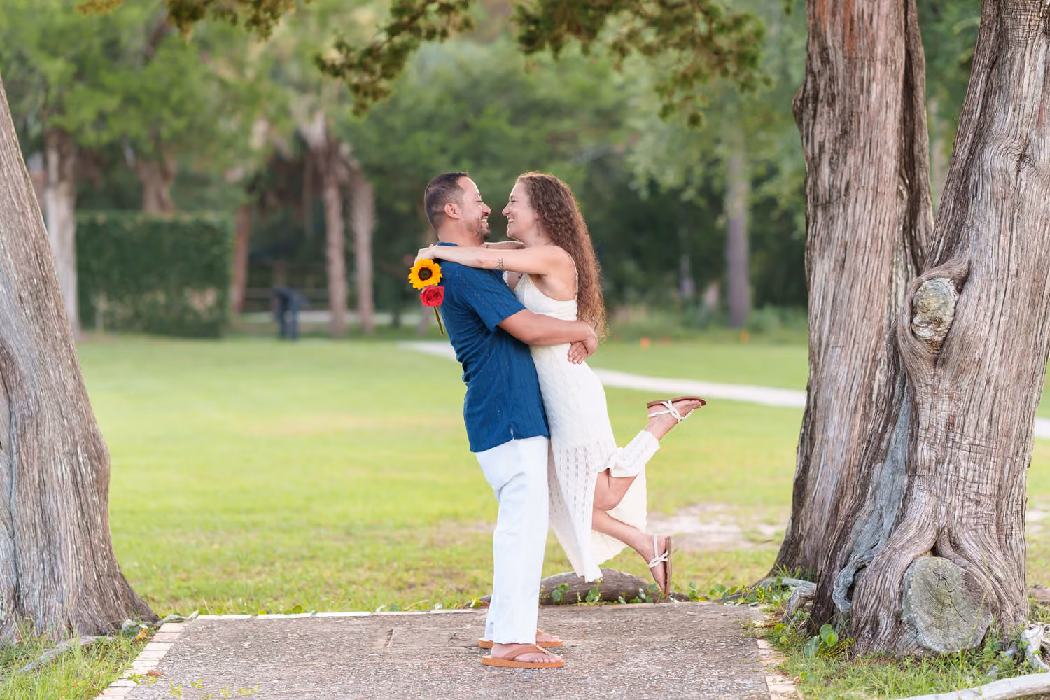 Woman holding sunflowers as her fiancé lifts her on a path between two large trees at Huntington Beach State Park