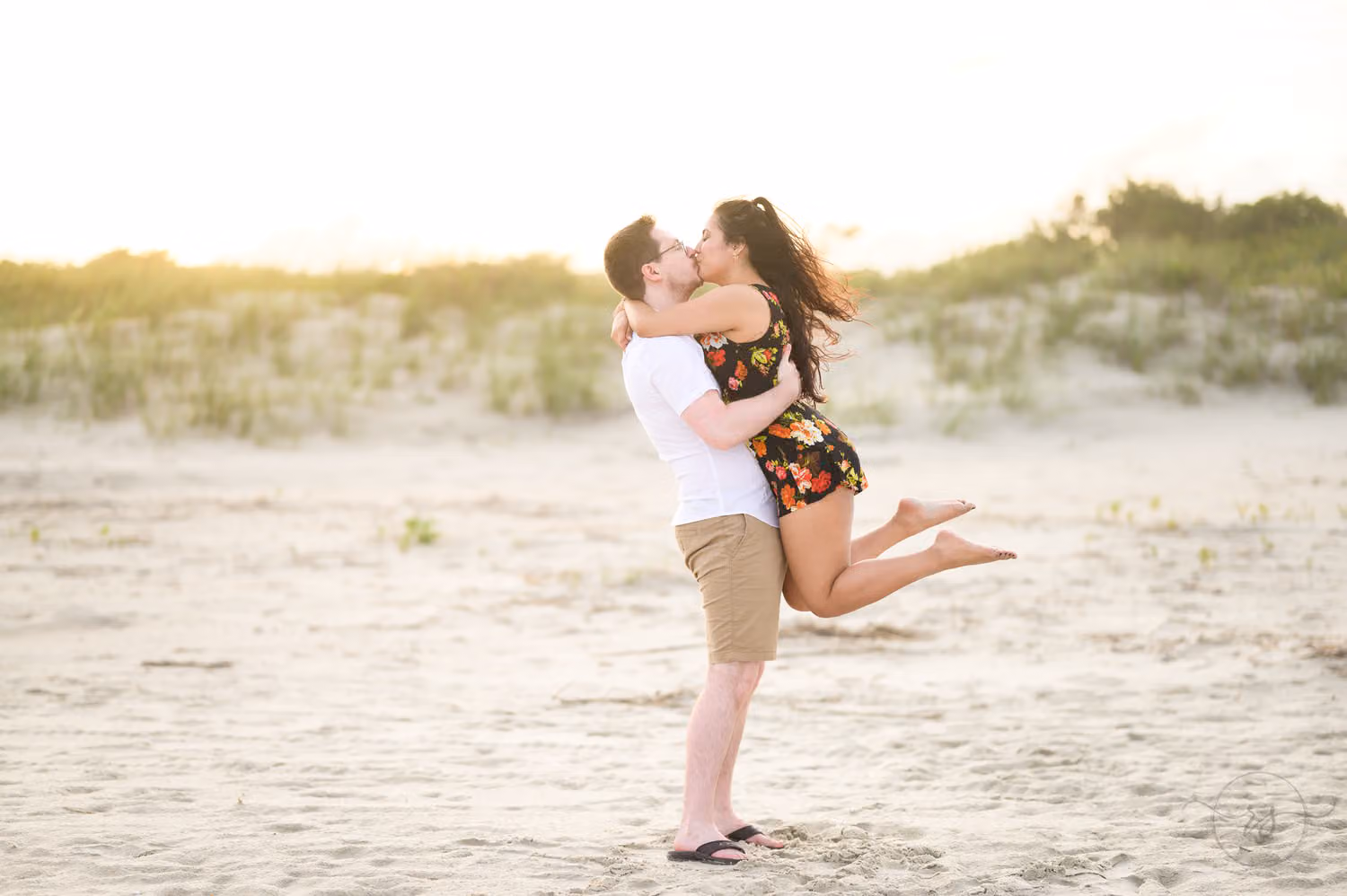 Man lifting his fiancée off the sand for a kiss as she kicks up her feet near the dunes at Huntington Beach State Park