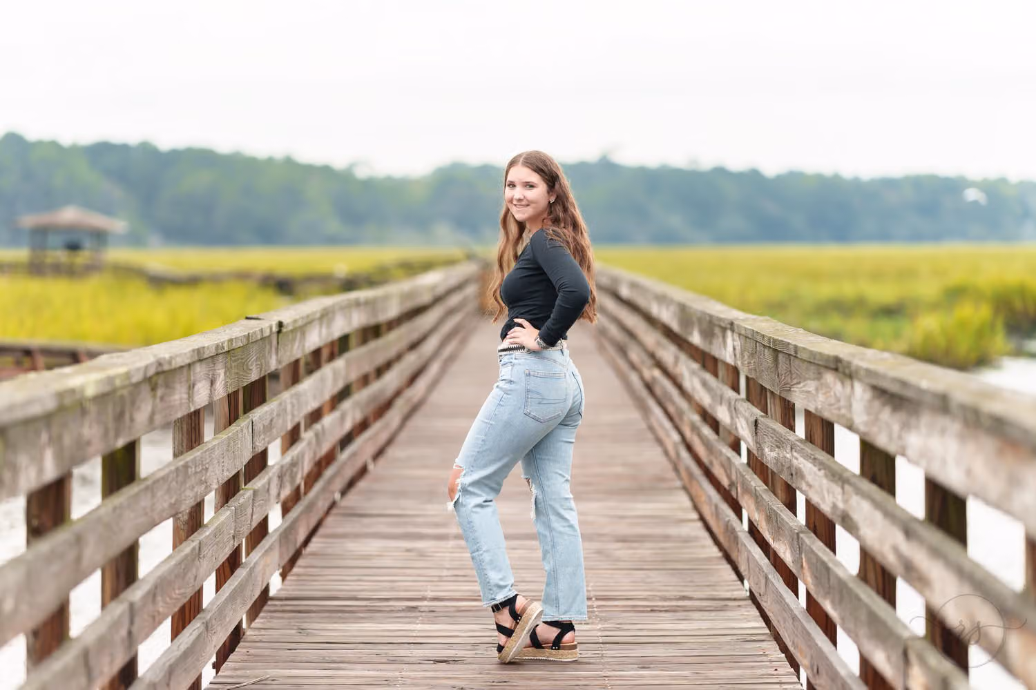 Senior in jeans standing on the marsh boardwalk and looking back over her shoulder at Huntington Beach State Park