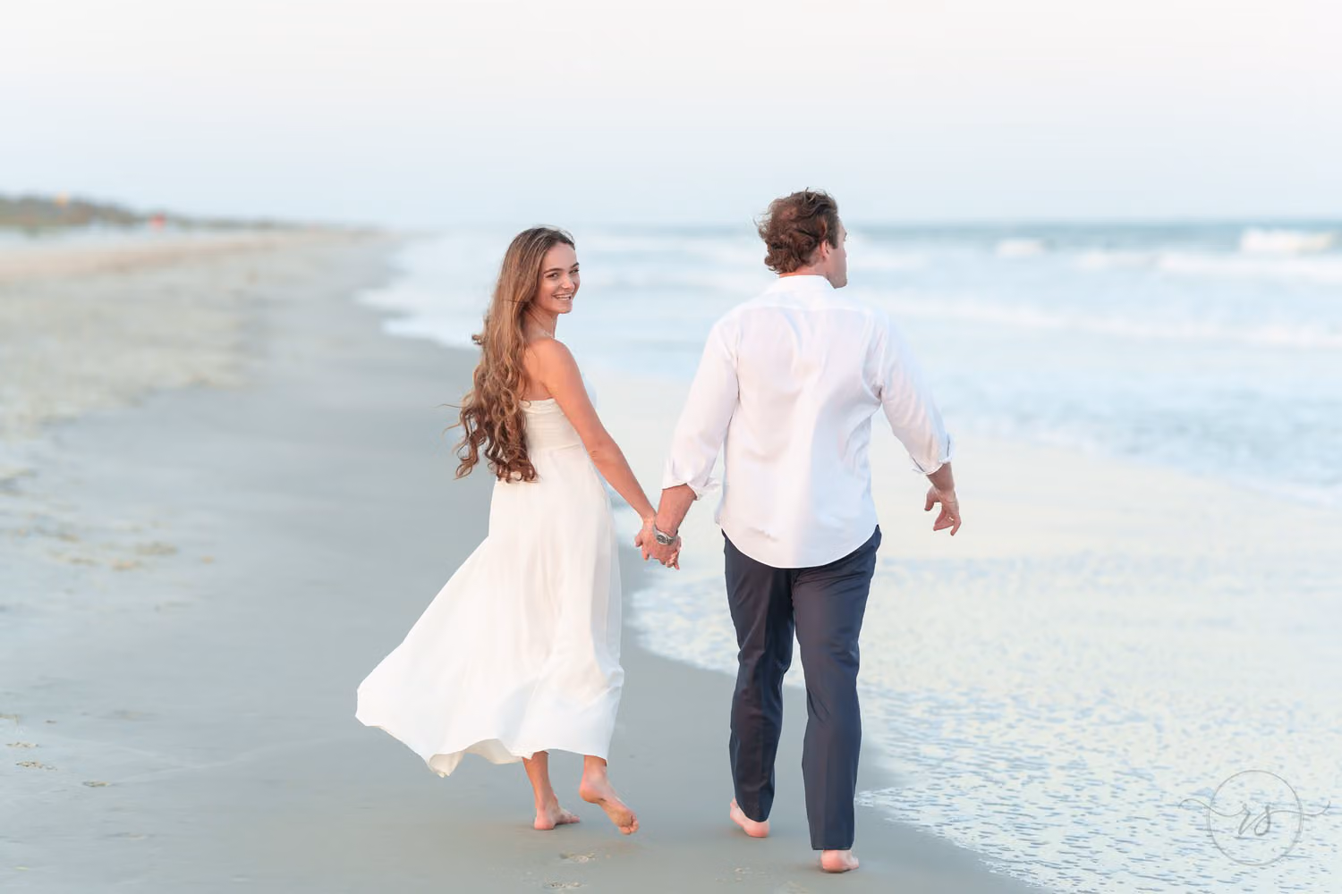 Woman in a white dress looking back as she walks hand in hand with her fiancé along the shore at Huntington Beach State Park