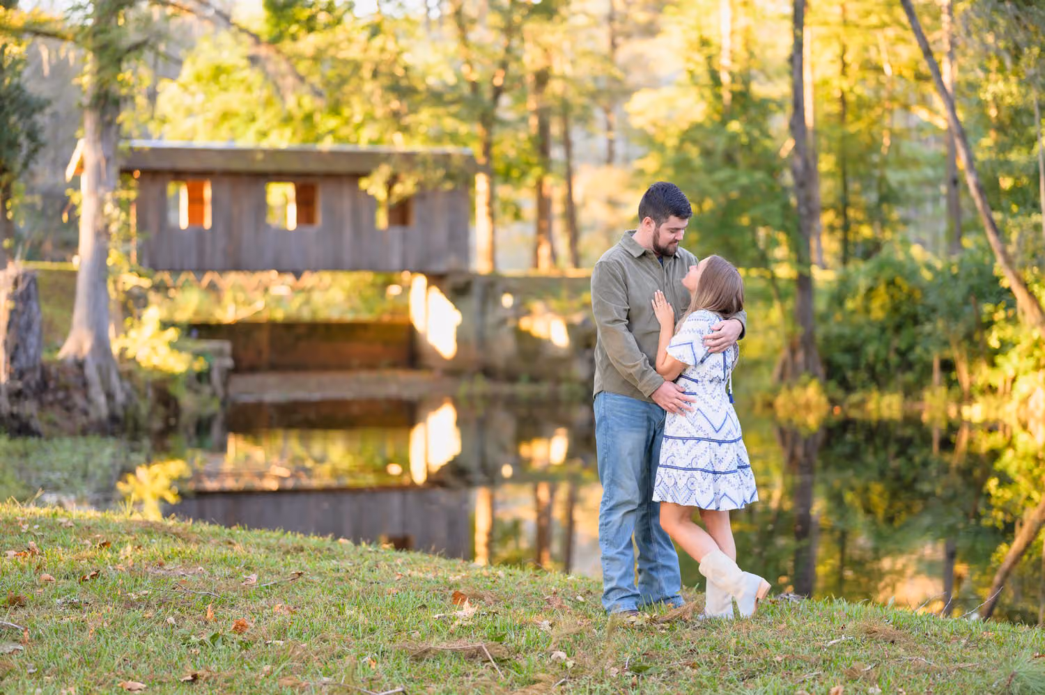 Couple smiling at each other on the grass with a covered bridge reflected in the pond behind them at Pine Log Plantation
