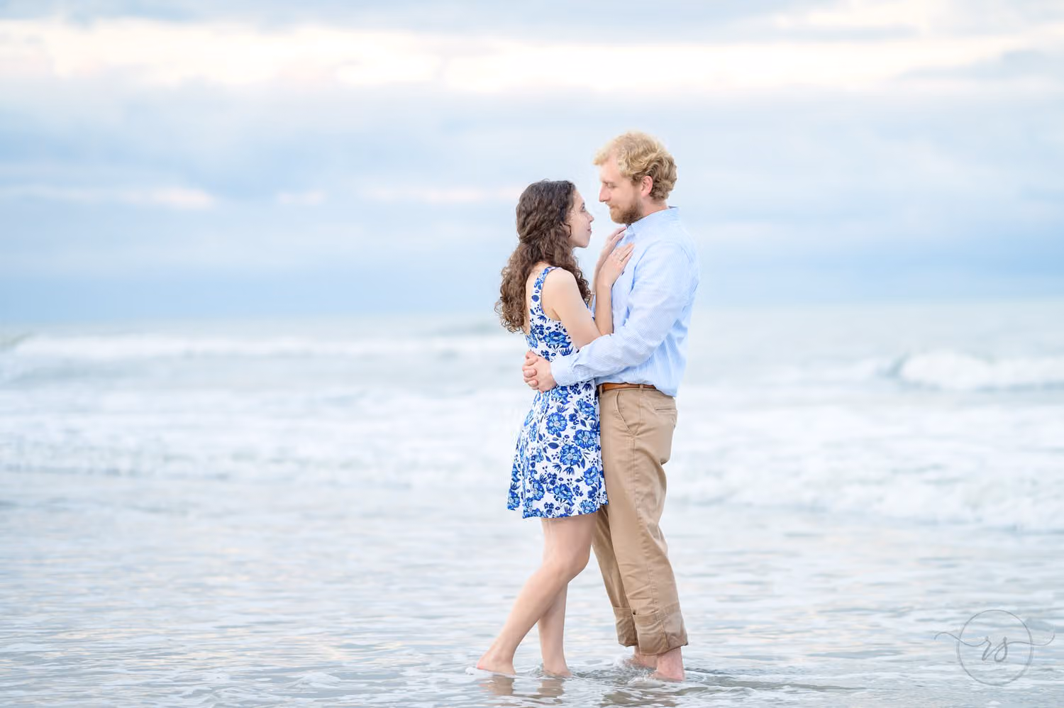 Couple standing ankle deep in the ocean gazing at each other under a soft overcast sky at Huntington Beach State Park