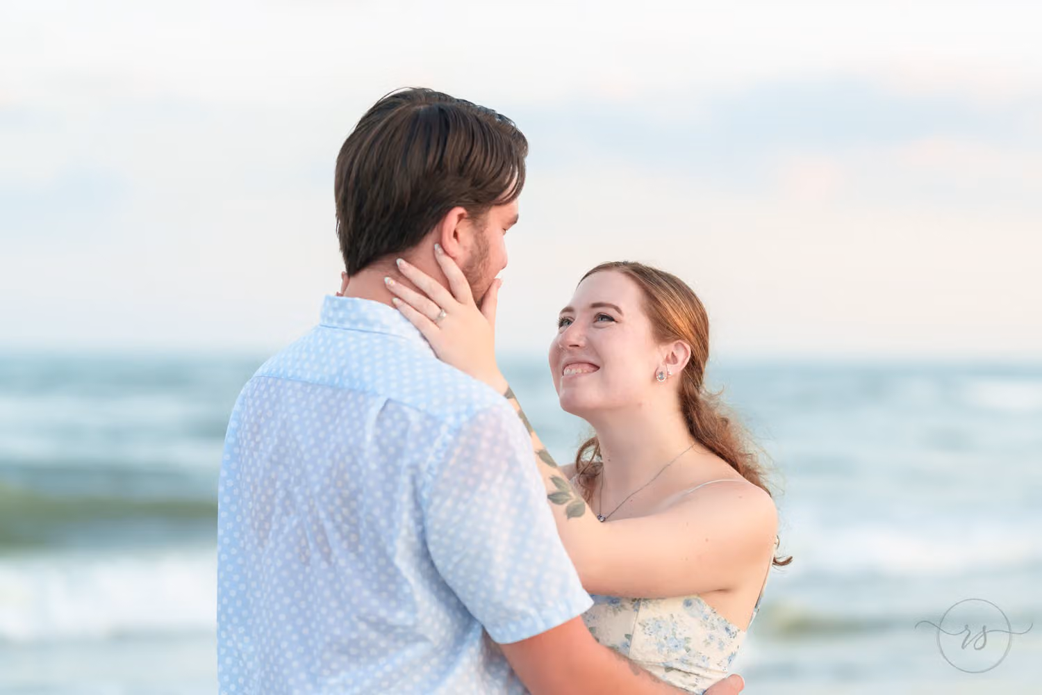 Woman smiling up at her fiancé as he holds her close by the ocean at Huntington Beach State Park