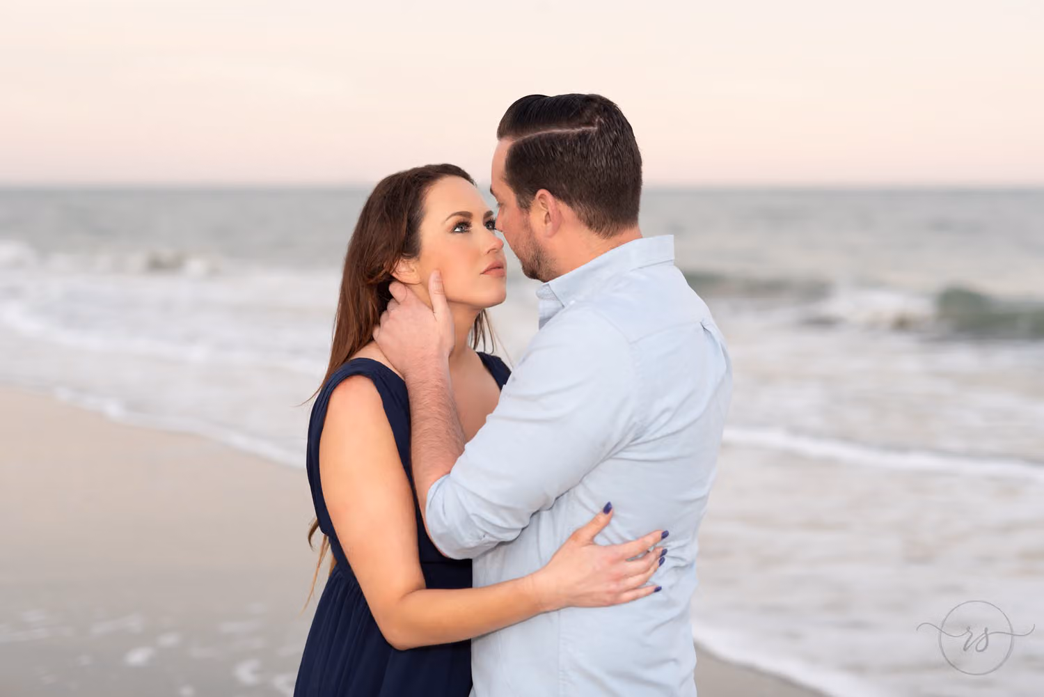 Man cupping his fiancée's face as she gazes up at him by the ocean at Huntington Beach State Park