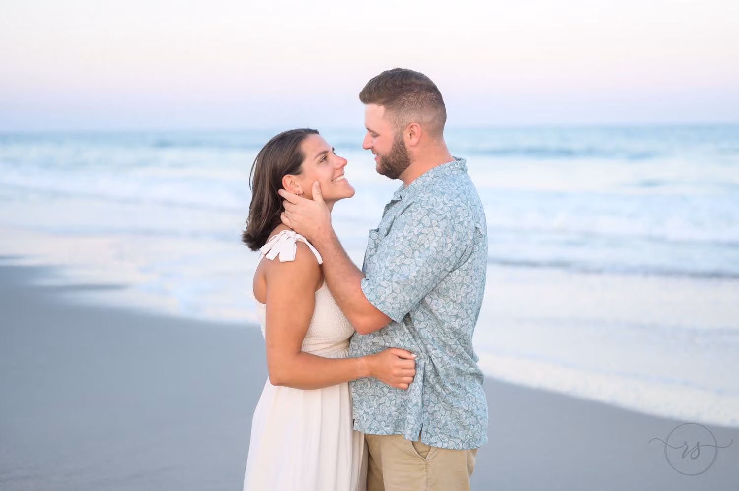 Man cradling the face of his fiancée in a white dress as they gaze at each other by the ocean at Huntington Beach State Park