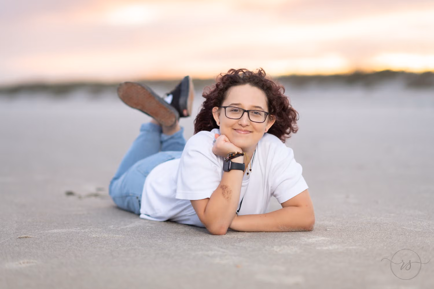 Senior in glasses lying on the sand with her chin on her hand and sneakers in the air at Huntington Beach State Park