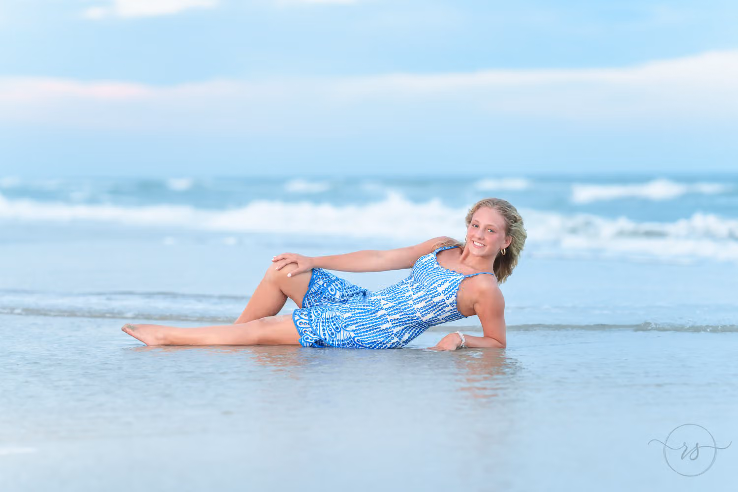 Senior girl in a blue patterned dress lying on her side in the shallow water at Huntington Beach State Park