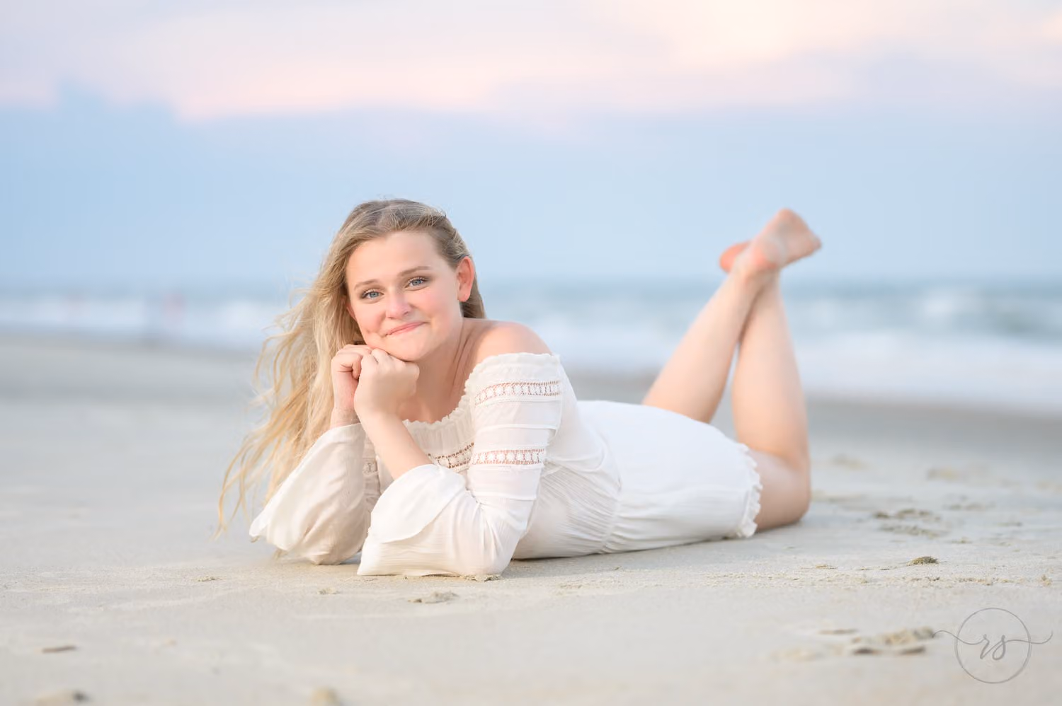 Smiling senior in a white lace dress lying on the sand with her feet kicked up at Huntington Beach State Park