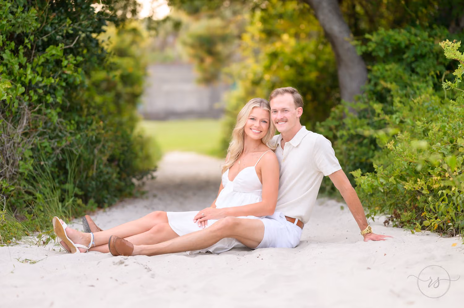 Couple in white reclining on a sandy path framed by green trees at Huntington Beach State Park