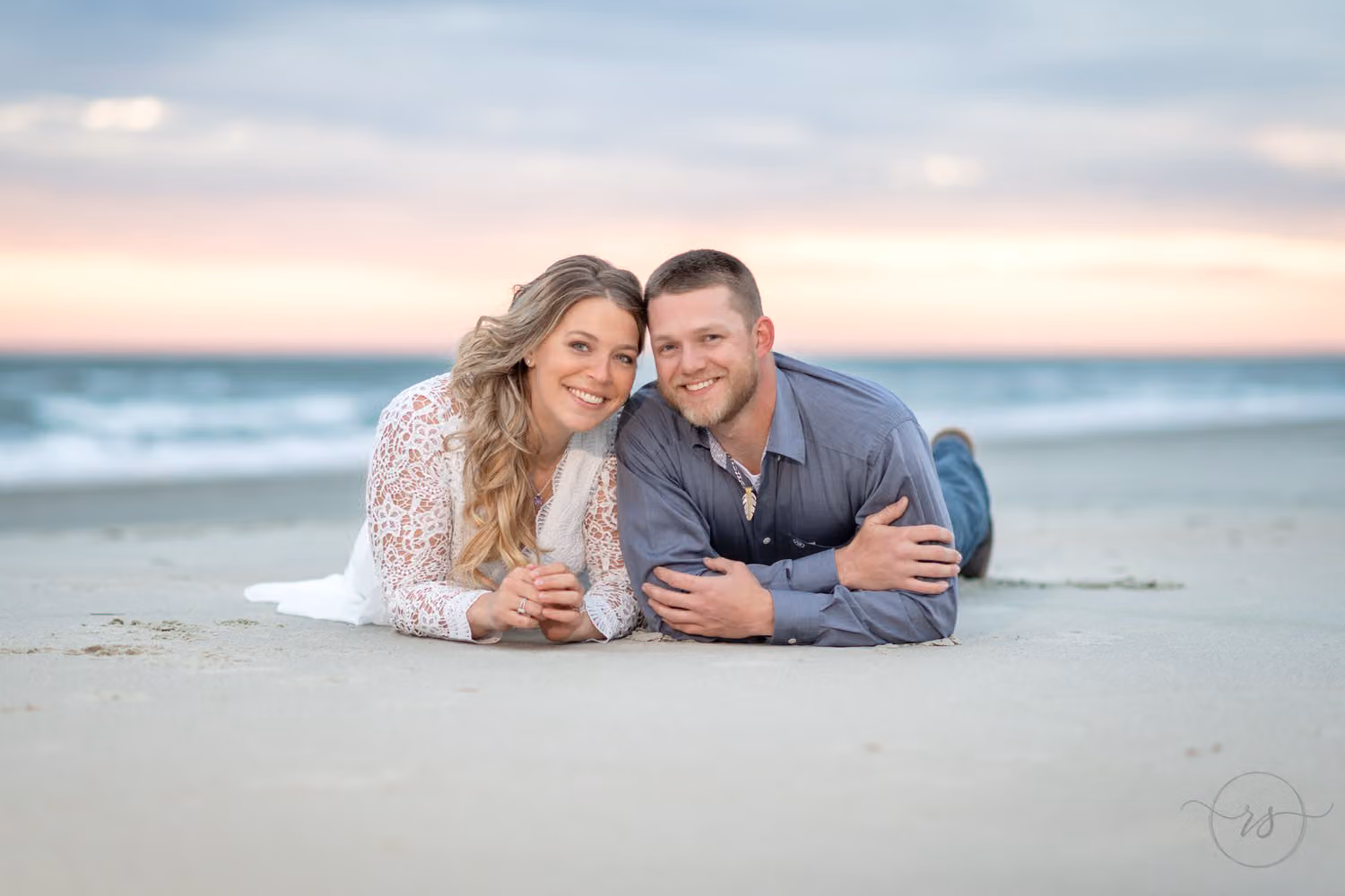 Couple lying side by side on the sand smiling, with waves and a pink sky behind them at Huntington Beach State Park
