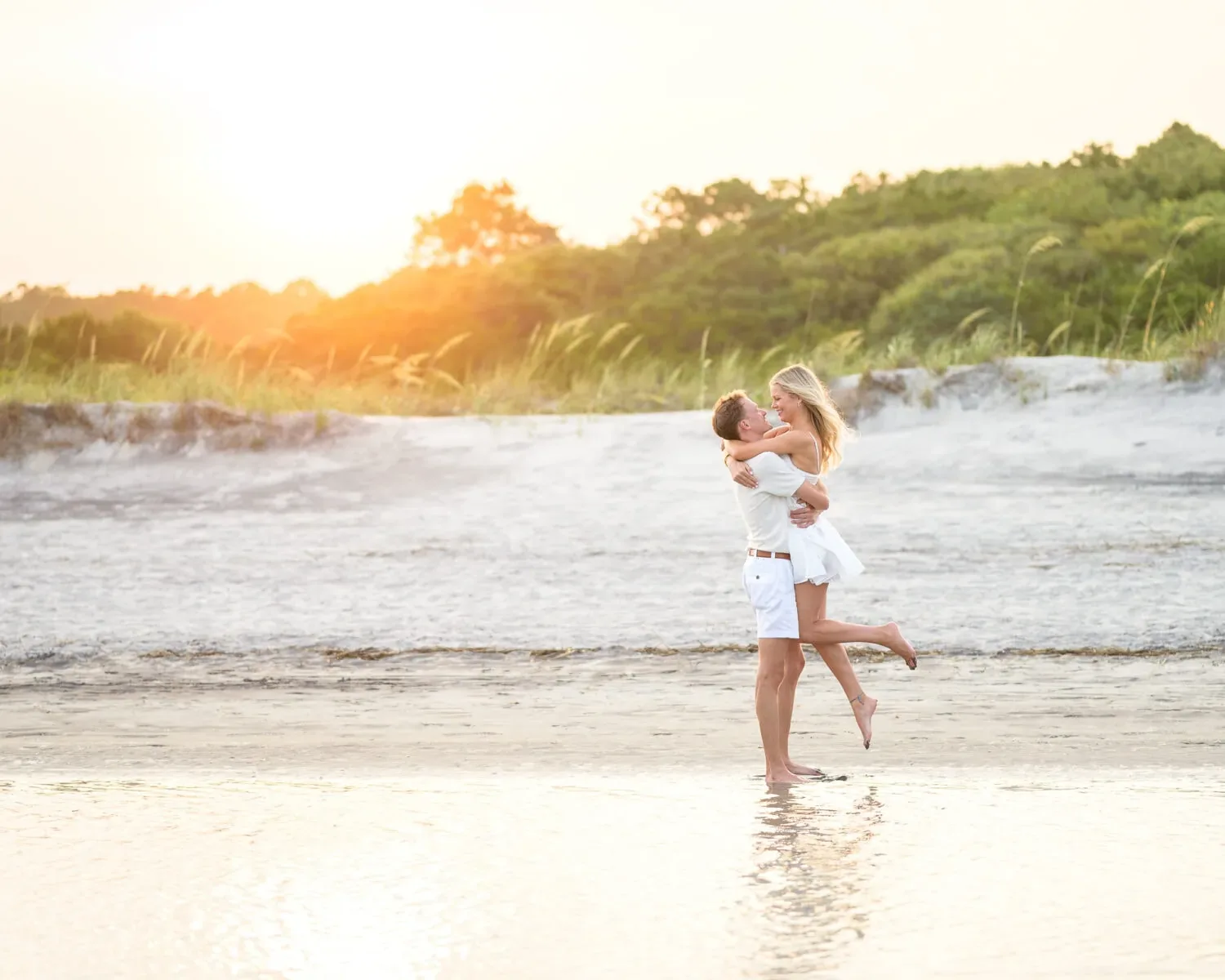 Man lifting his partner off the sand at the water's edge with the sunset behind them at Huntington Beach State Park