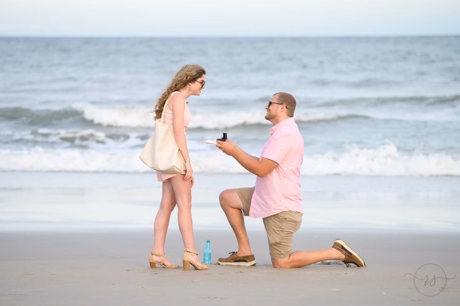 Man on one knee holding up a ring beside a message bottle as she reacts at the water's edge at Huntington Beach State Park