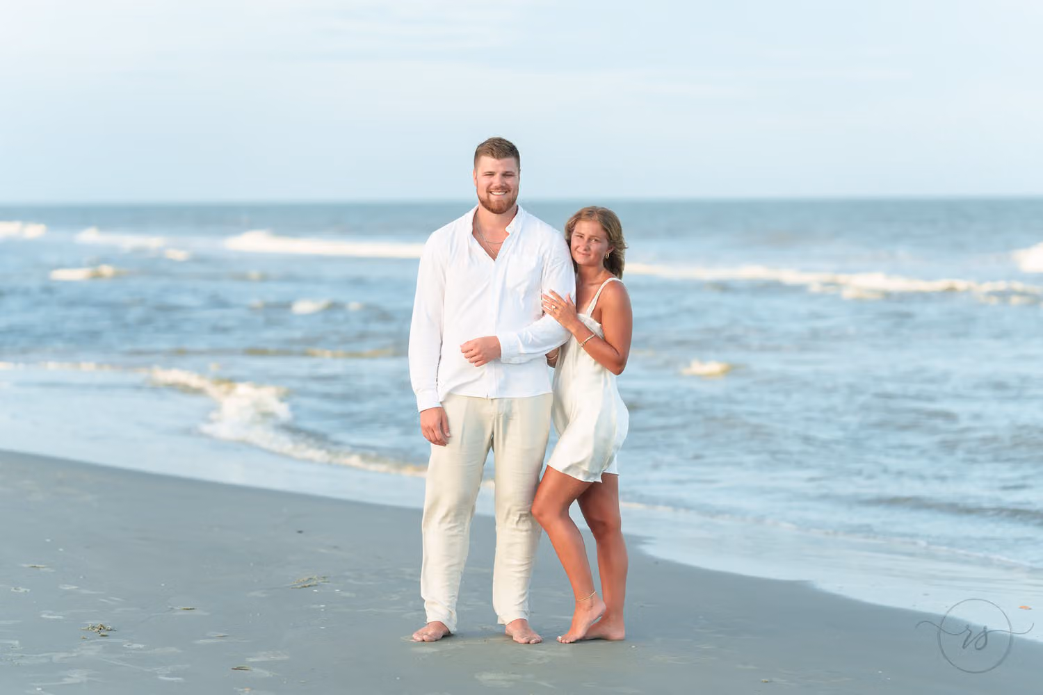 Man in white and his fiancée in a white dress smiling side by side at the water's edge in Pawleys Island