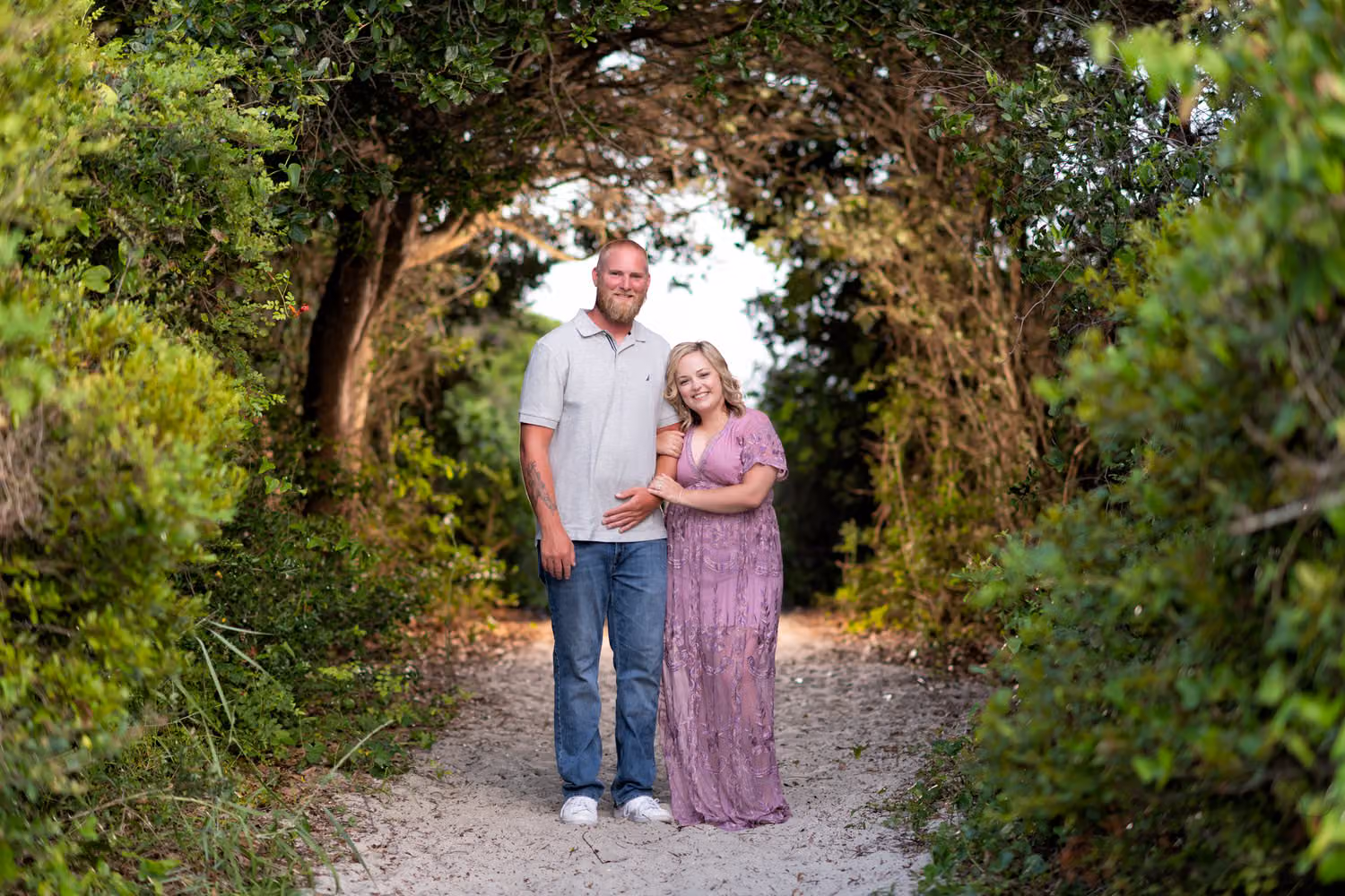 Couple standing close at the end of a sandy path framed by an arch of trees at Huntington Beach State Park