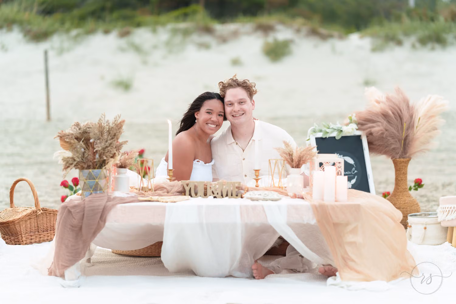 Couple sitting at a low picnic table set with candles, pampas grass and a sign on the sand at Myrtle Beach State Park
