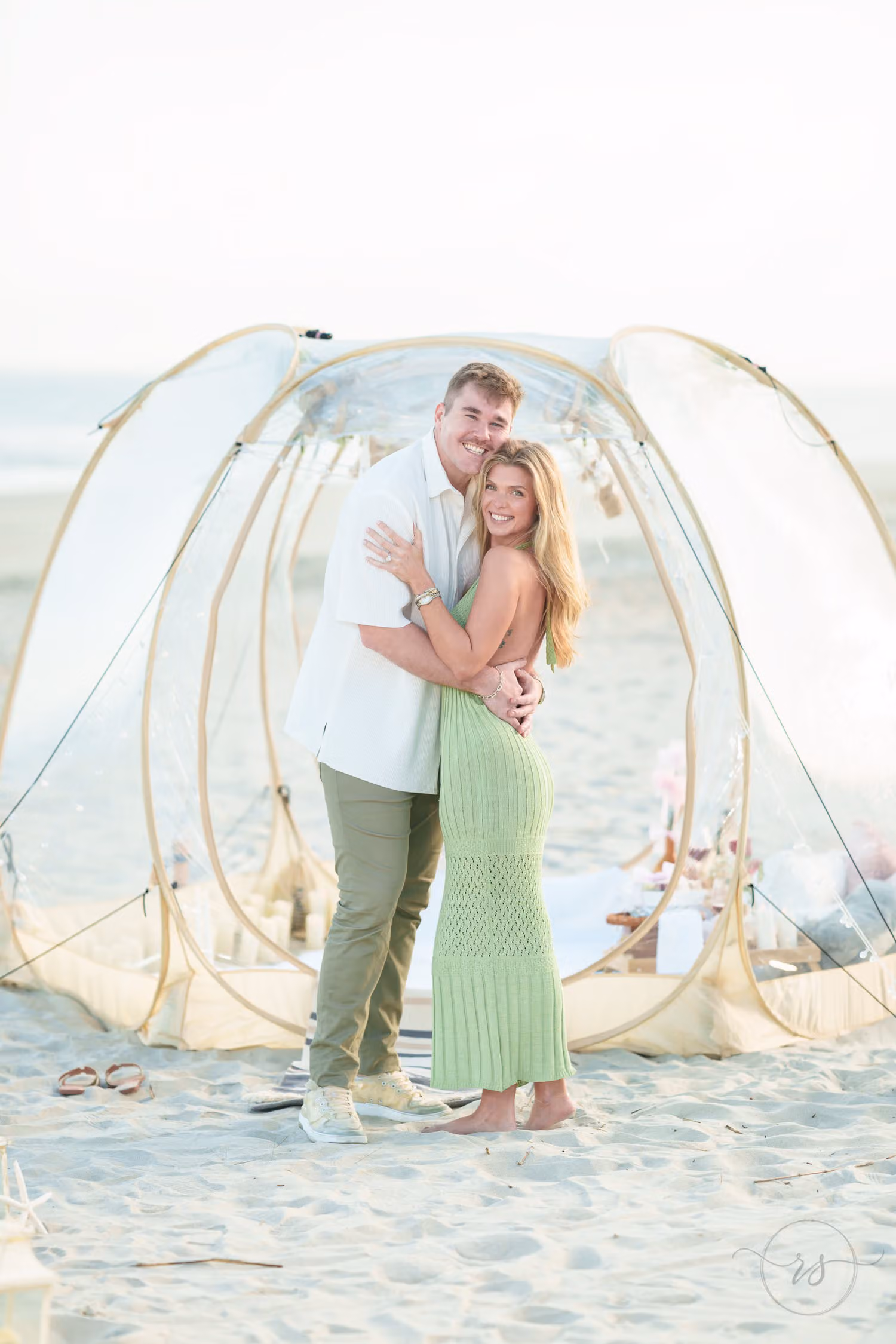 Couple standing in an embrace inside a clear bubble tent set up for a beach picnic at Huntington Beach State Park