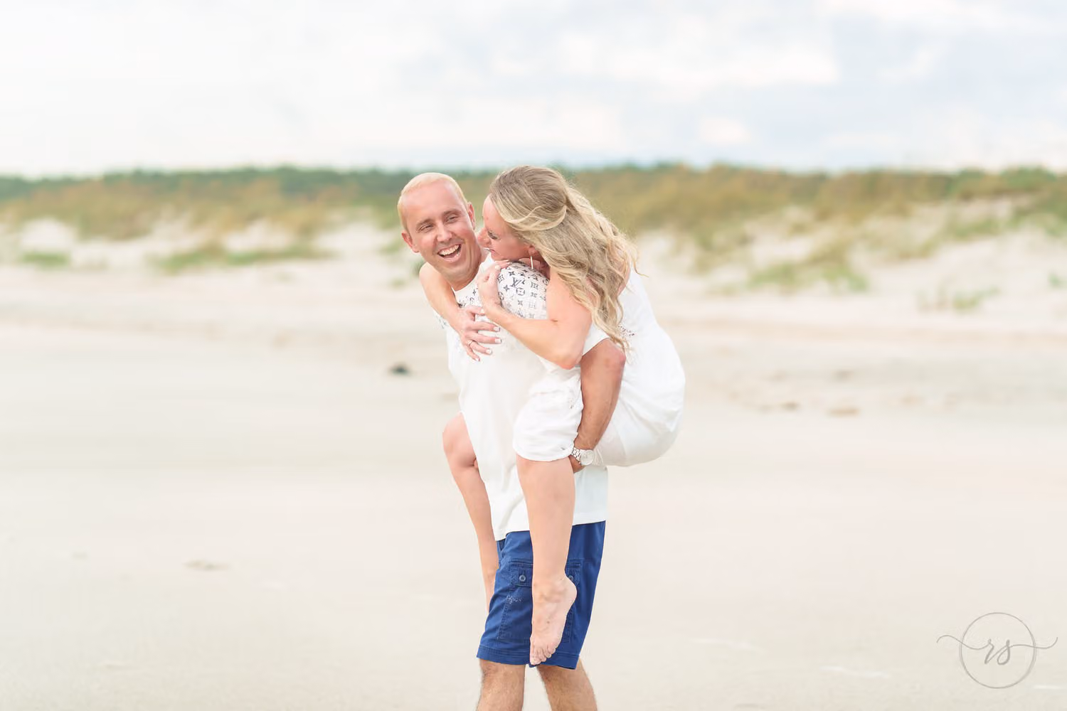 Woman riding piggyback and kissing her laughing fiancé on the cheek on the beach by the dunes at Huntington Beach State Park