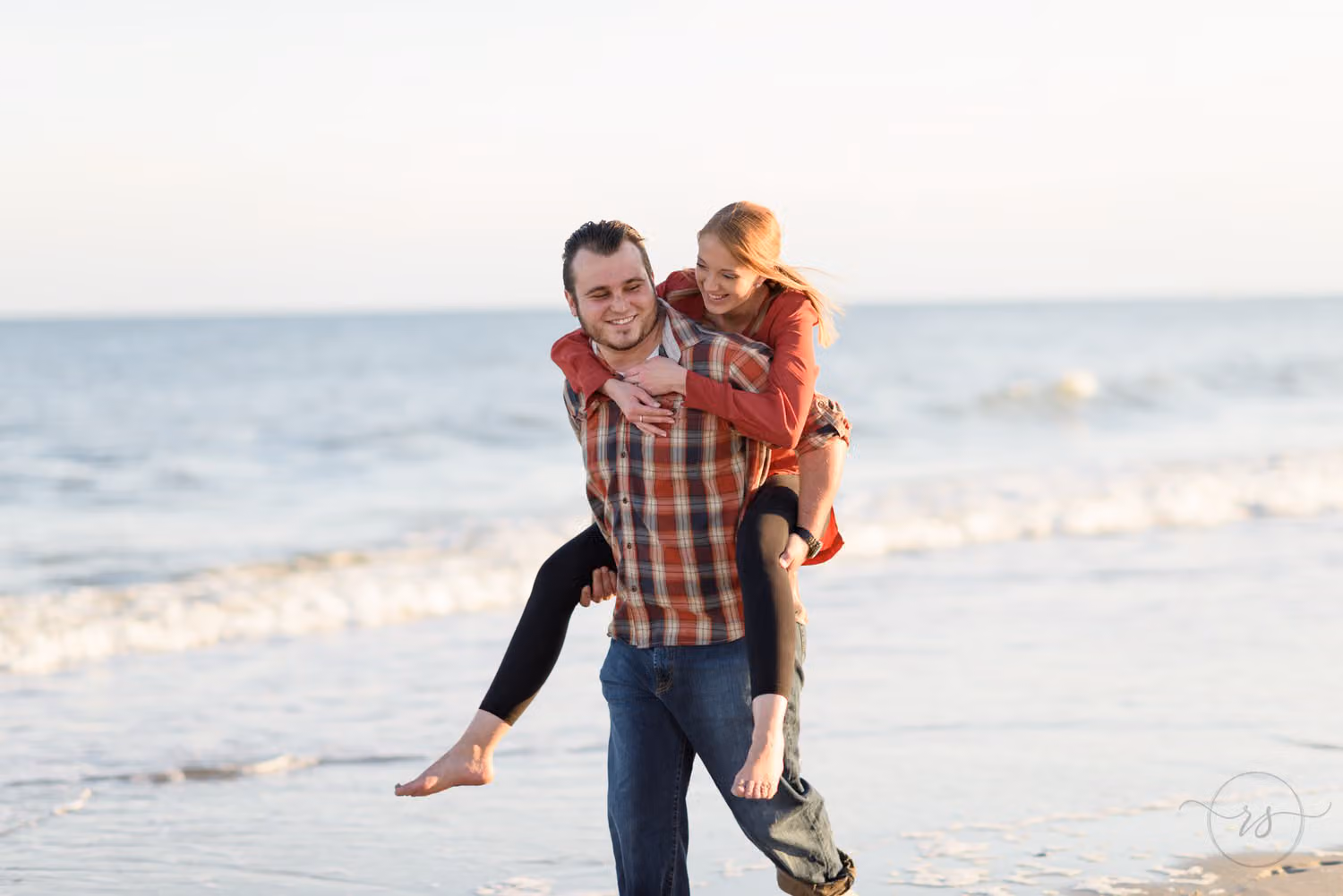 Woman riding piggyback on her fiancé, both laughing, along the water's edge at Myrtle Beach State Park