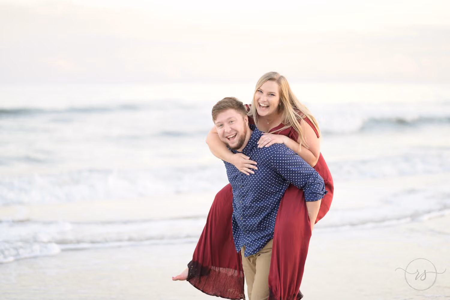 Laughing woman in a red dress riding piggyback on her fiancé at the edge of the surf at Huntington Beach State Park