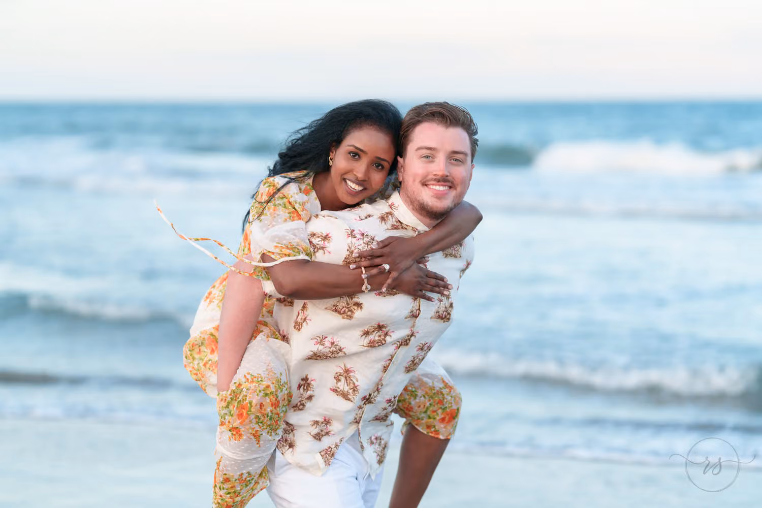 Laughing woman riding piggyback on her fiancé in a floral shirt by the waves in Pawleys Island