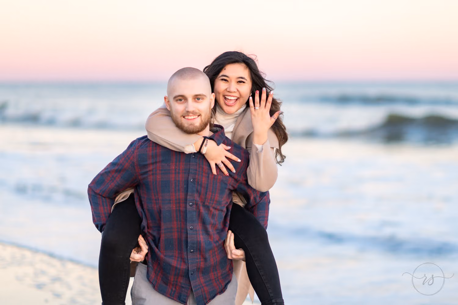 Woman laughing on a piggyback ride and holding up her ring under a pink sky by the ocean at Huntington Beach State Park