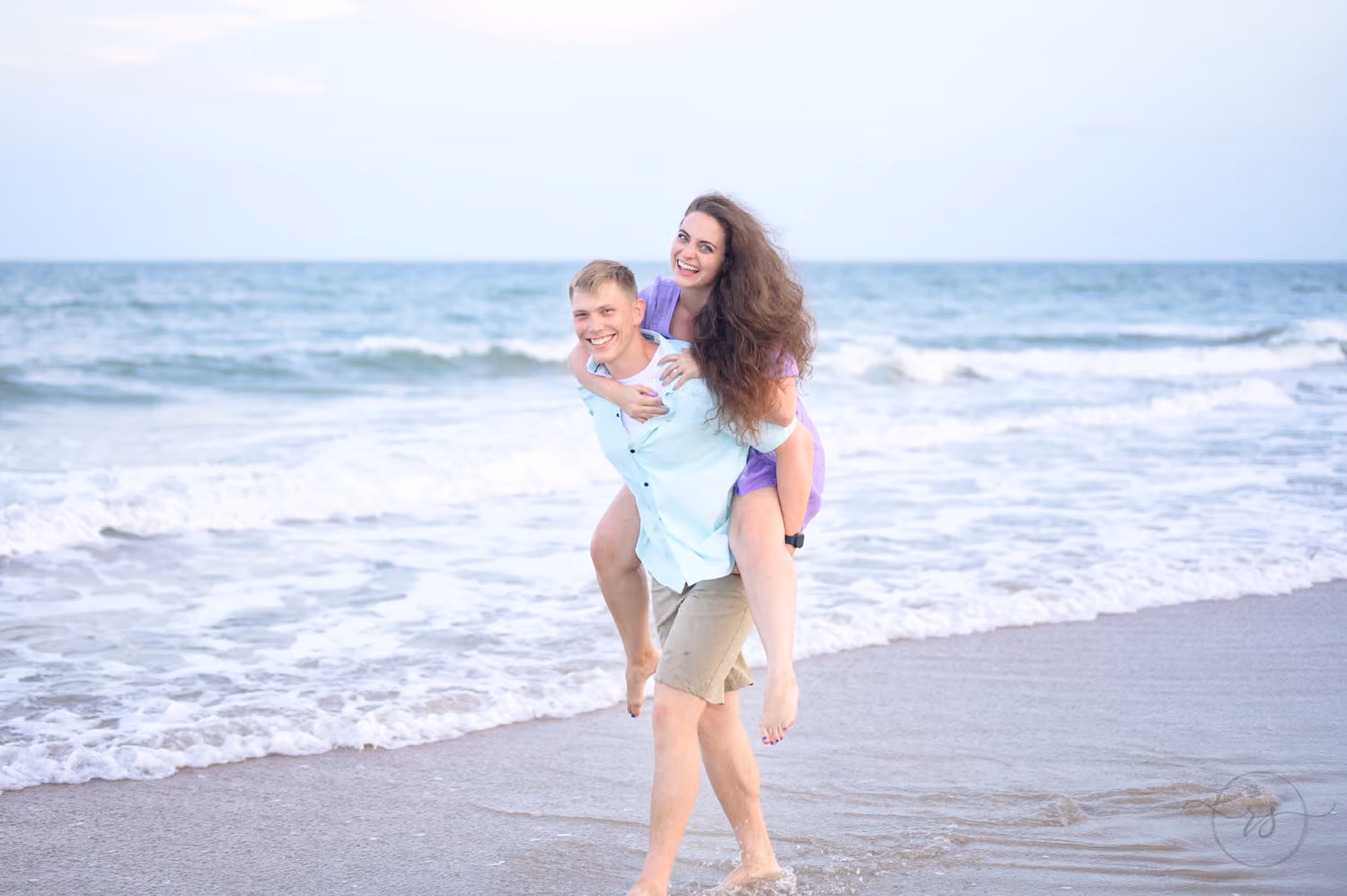Woman riding piggyback on her fiancé as he walks through the shallow surf at Huntington Beach State Park