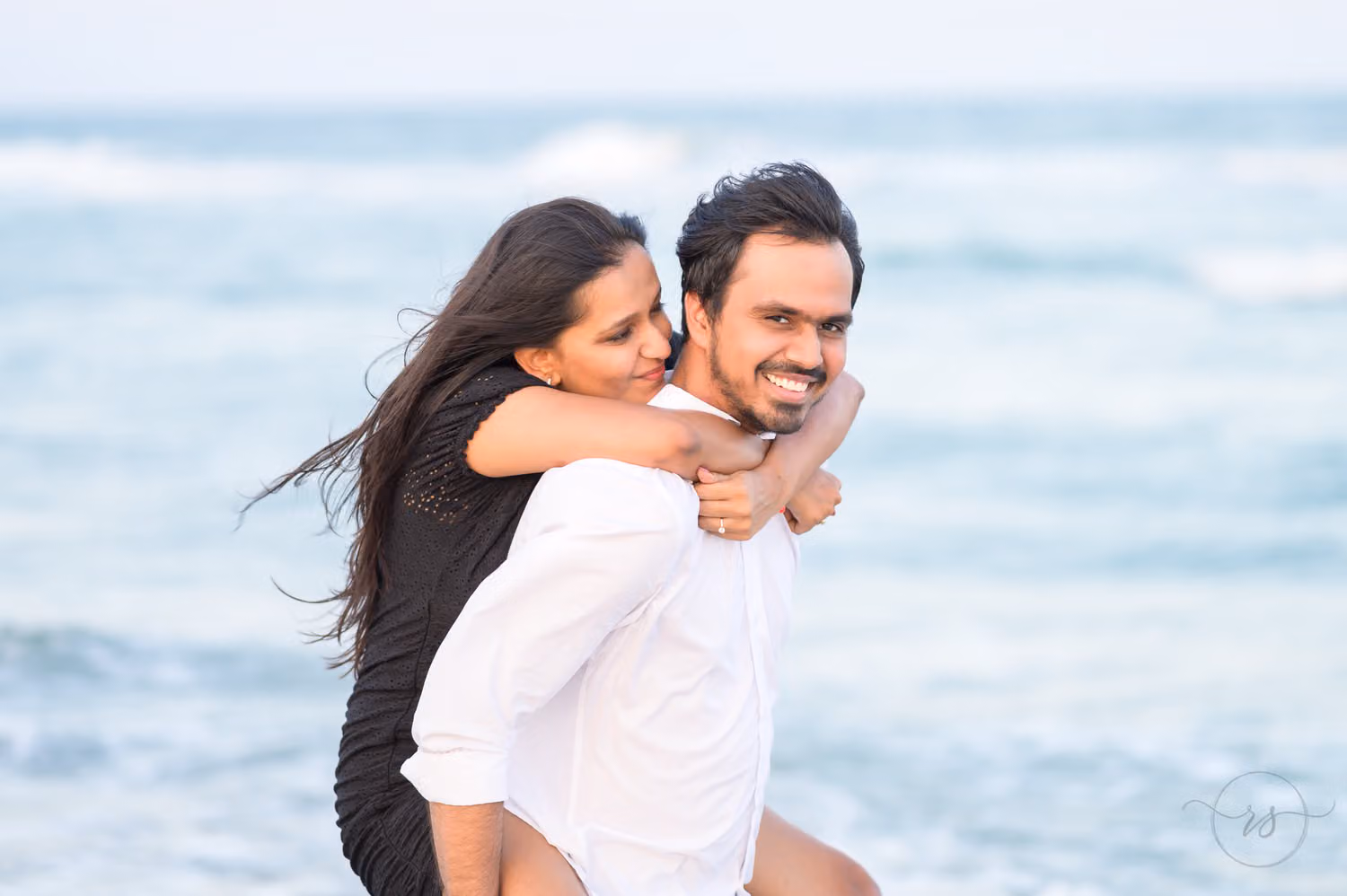 Woman with windblown hair riding piggyback on her smiling fiancé by the ocean at Huntington Beach State Park