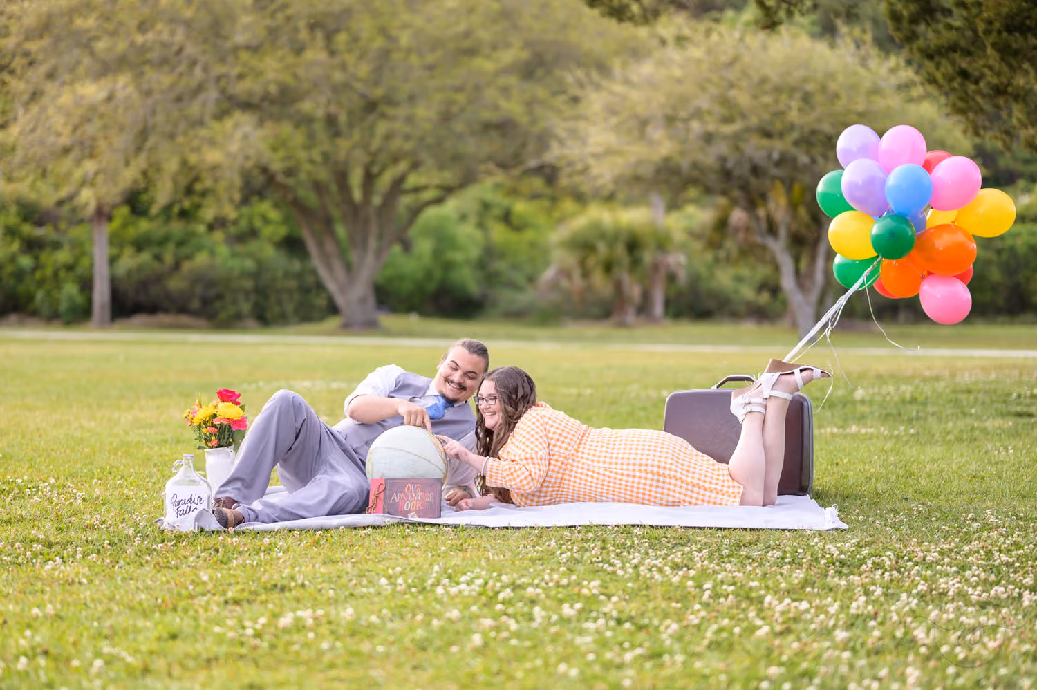 Couple on a picnic blanket in the grass with colorful balloons and an Up-style suitcase at Huntington Beach State Park