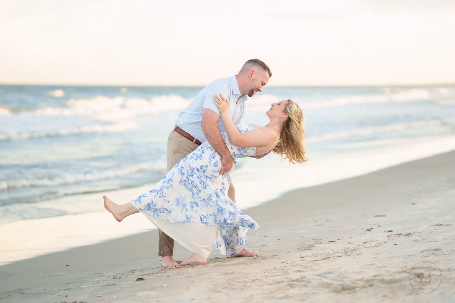 Man dipping his fiancée in a blue floral dress as she kicks up her foot on the beach at Huntington Beach State Park