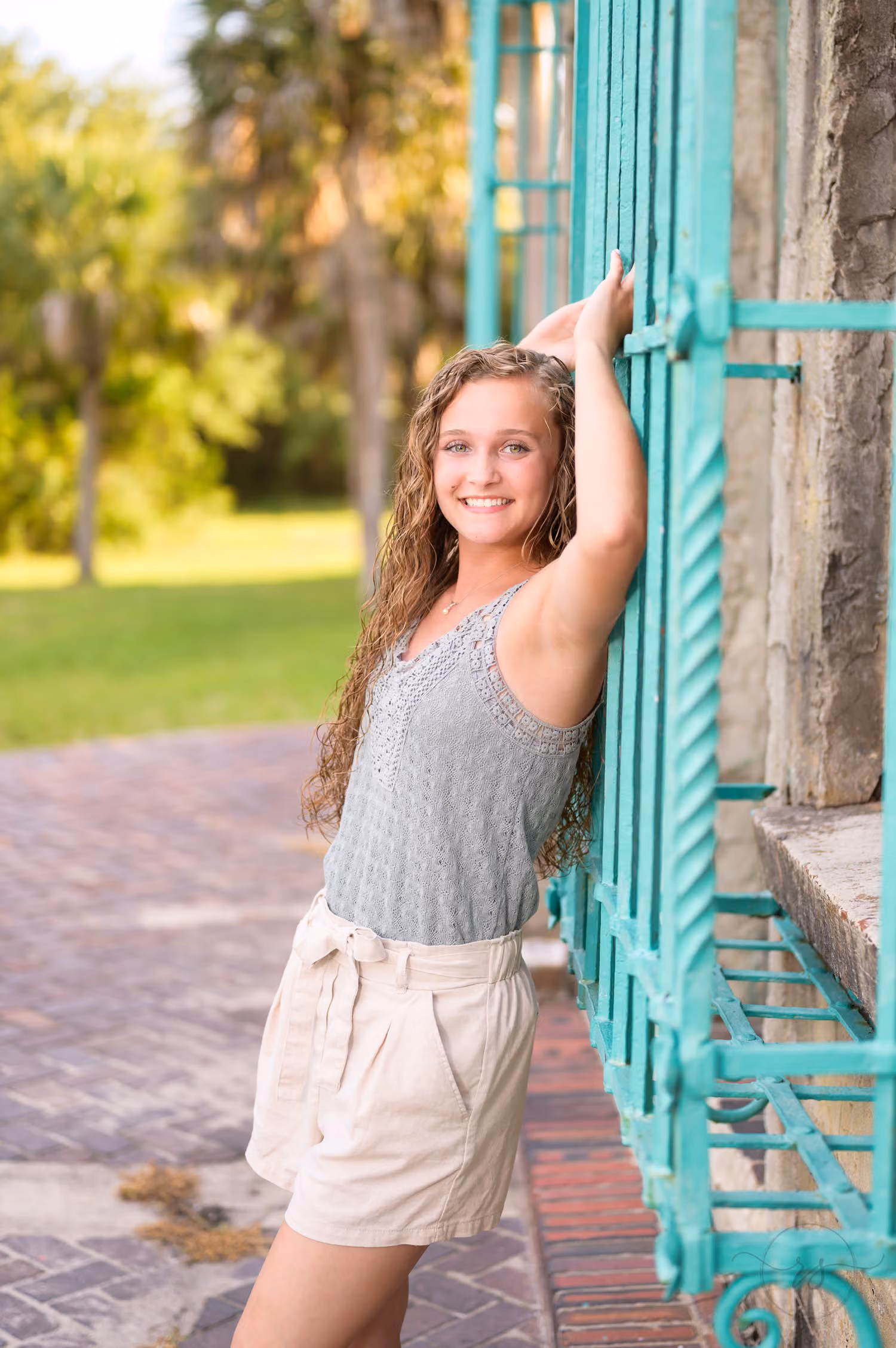 Senior girl in a gray top and shorts leaning on the teal iron window grille at Atalaya Castle at Huntington Beach State Park