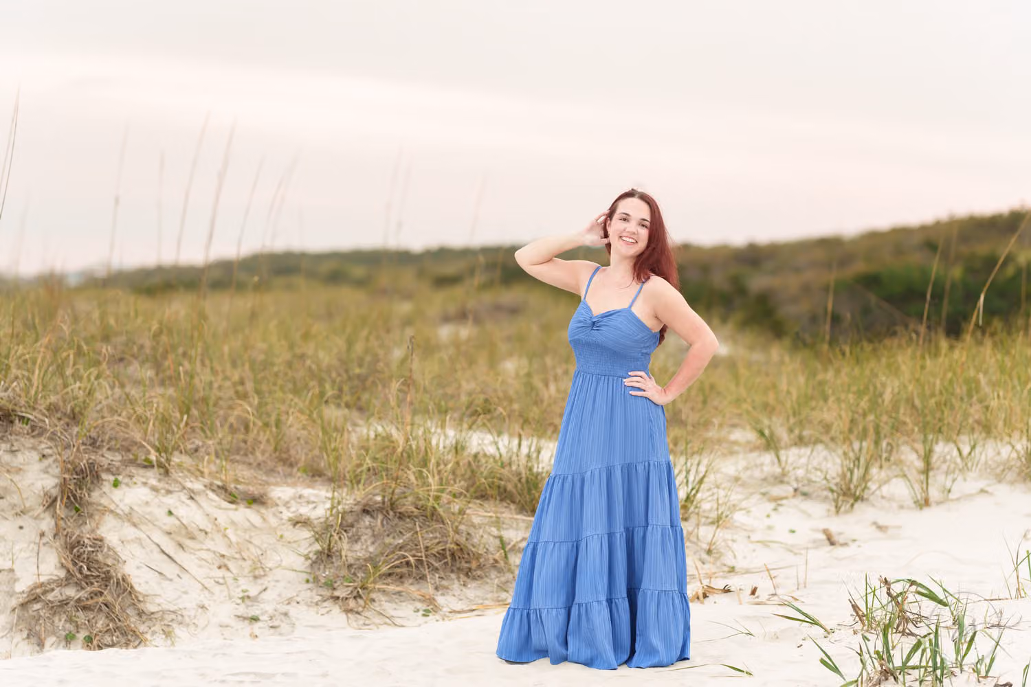 Red-haired senior in a long blue dress standing on the dunes under a soft cloudy sky at Huntington Beach State Park