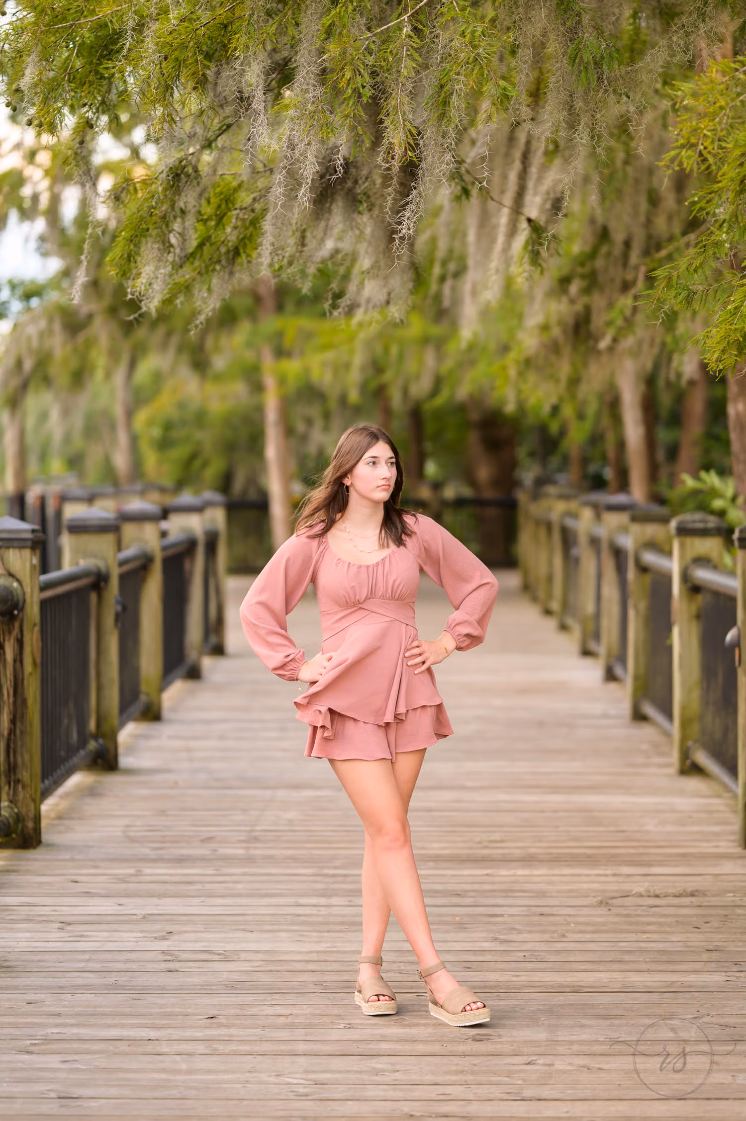 Senior girl in a pink dress standing on the boardwalk under oaks draped with Spanish moss at the Conway River Walk
