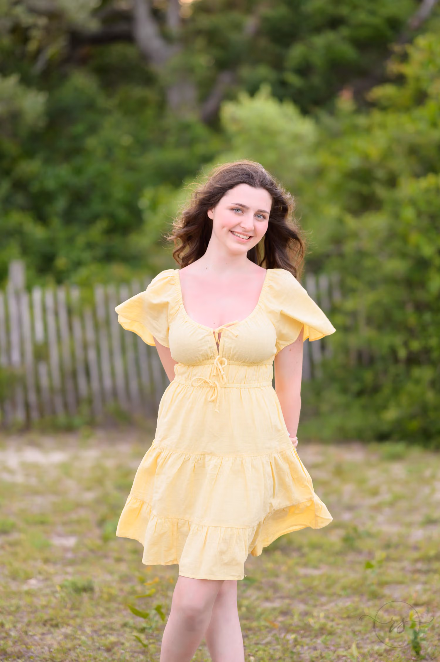 Senior in a yellow dress standing in front of a beach fence and green trees at Myrtle Beach State Park