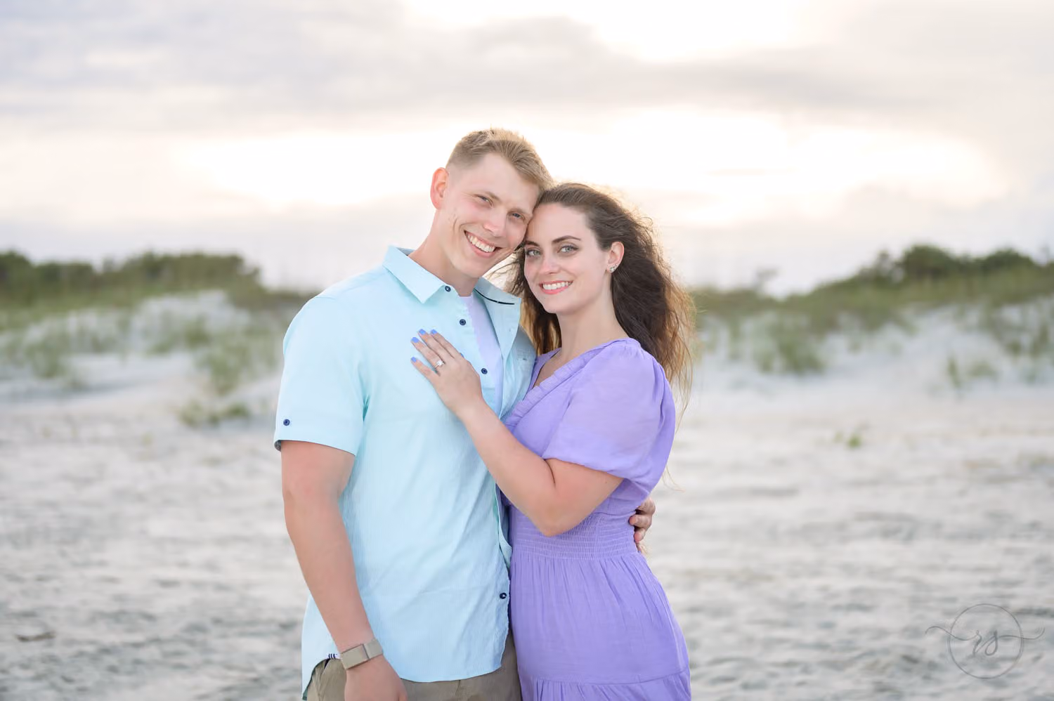 Couple hugging and smiling at the camera with the dunes behind them in evening light at Huntington Beach State Park