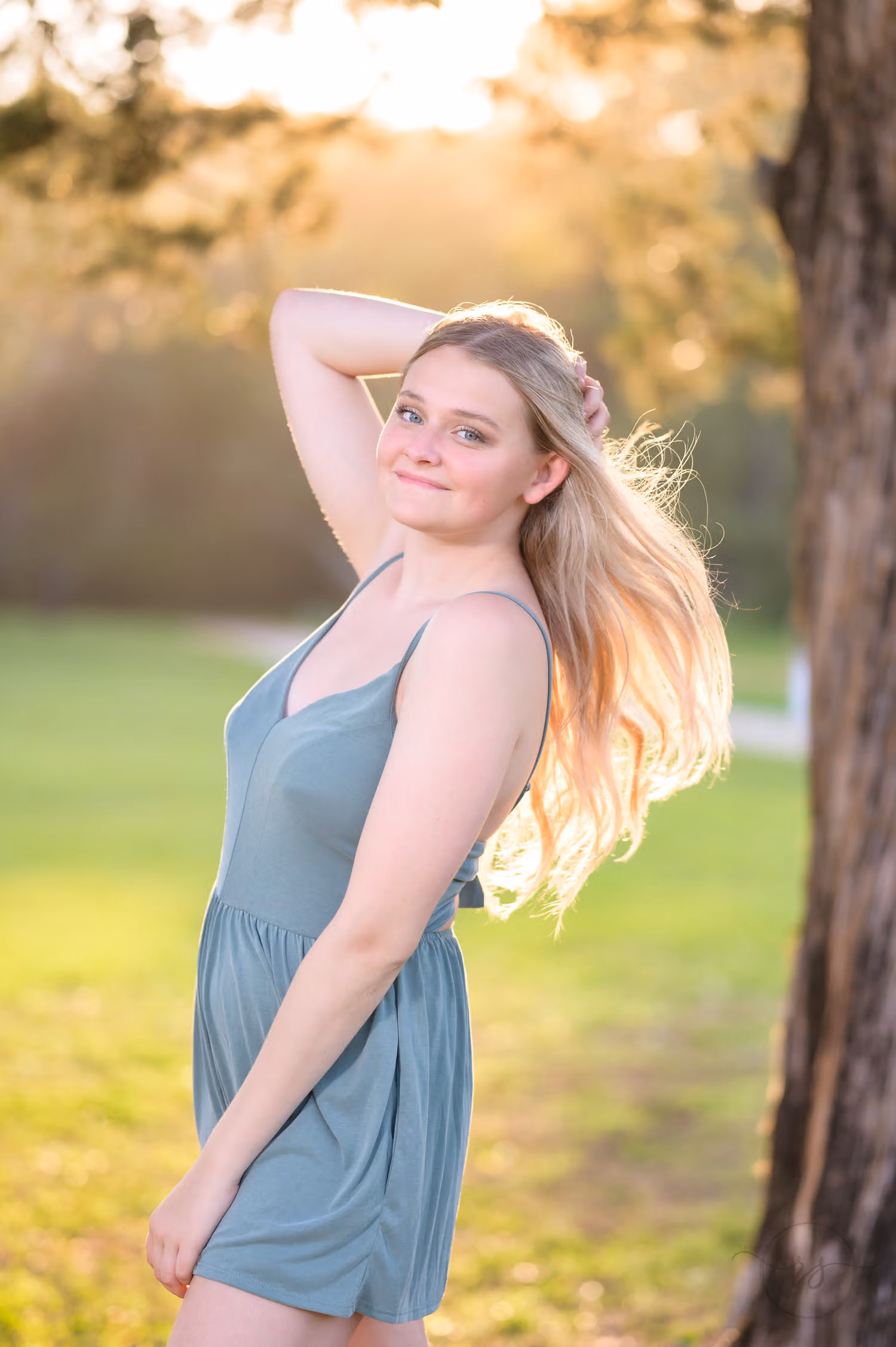Senior in a green romper with her hand in her hair, glowing in sunset backlight on the lawn at Huntington Beach State Park