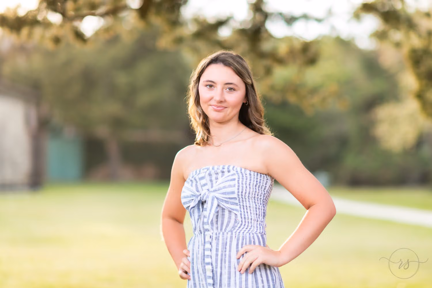 Senior in a striped strapless dress with a hand on her hip on a sunny lawn at sunset at Huntington Beach State Park