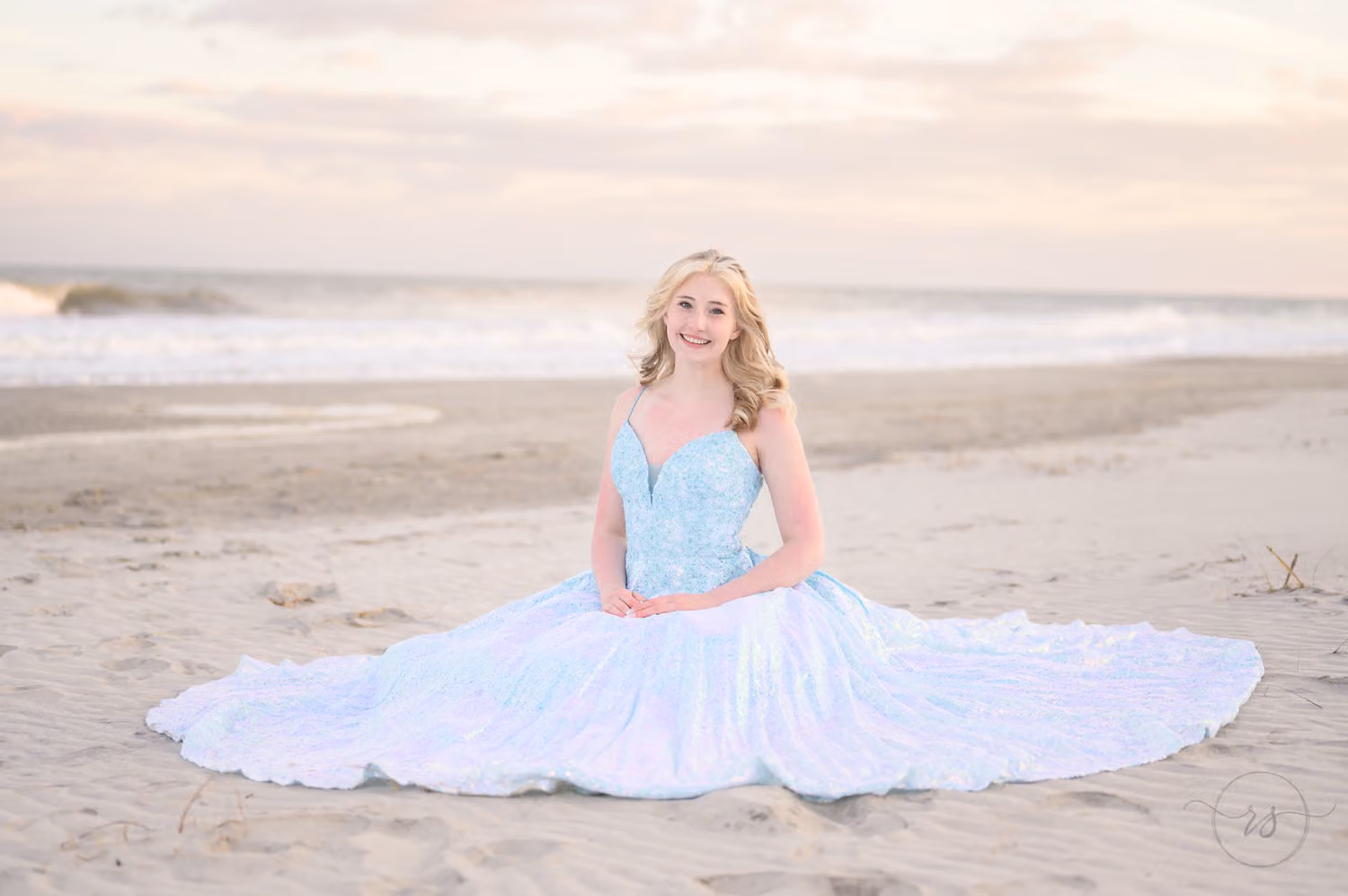 Teen sitting on the sand with her sparkly blue prom dress spread in a circle around her at Huntington Beach State Park