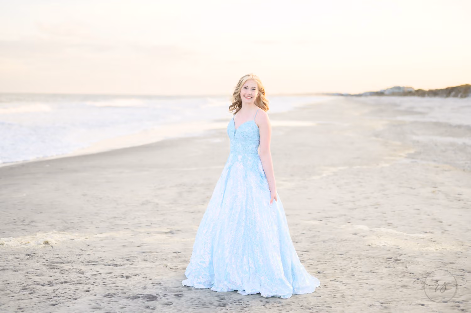Teen in a flowing blue prom gown standing on a wide stretch of beach with the ocean behind at Huntington Beach State Park
